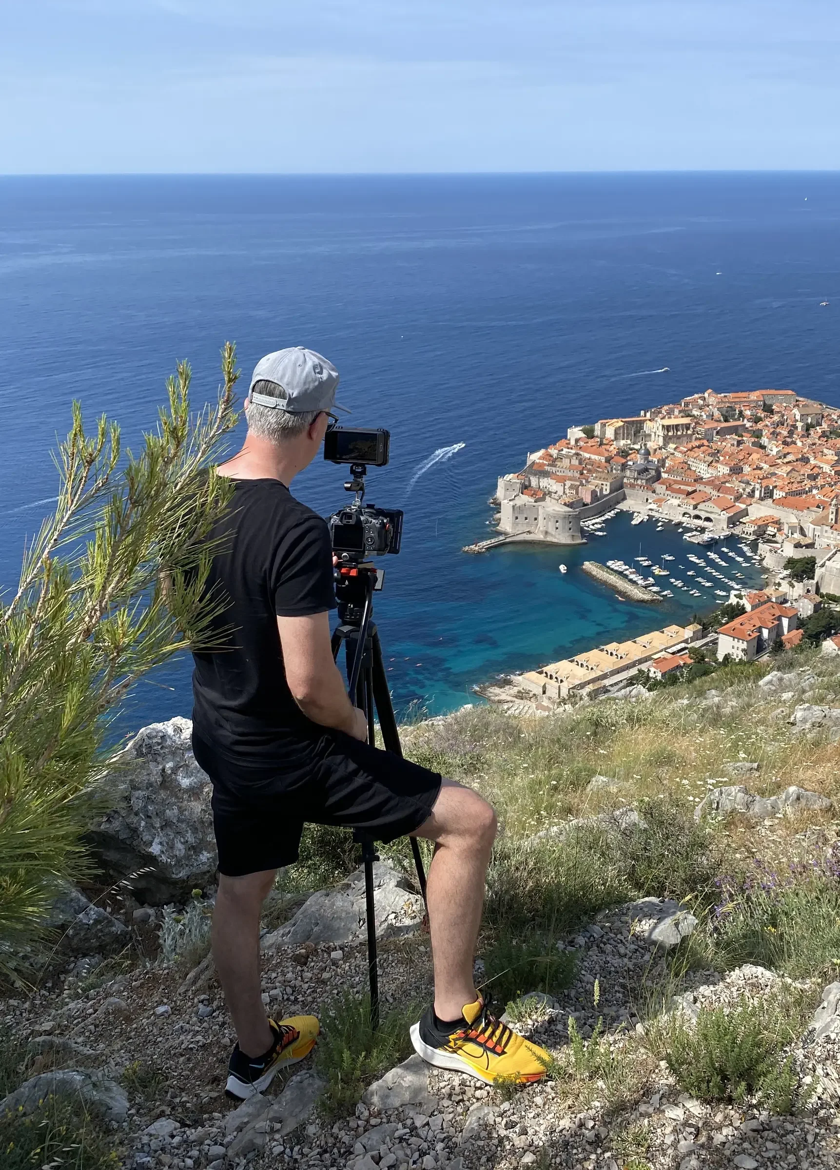 A man in black shorts, a black t-shirt, yellow sneakers, and a gray cap is standing on a rocky hill taking photos or videos of a coastal town with a camera on a tripod. The town has red-roofed buildings, a marina, and a city wall, with blue ocean wat