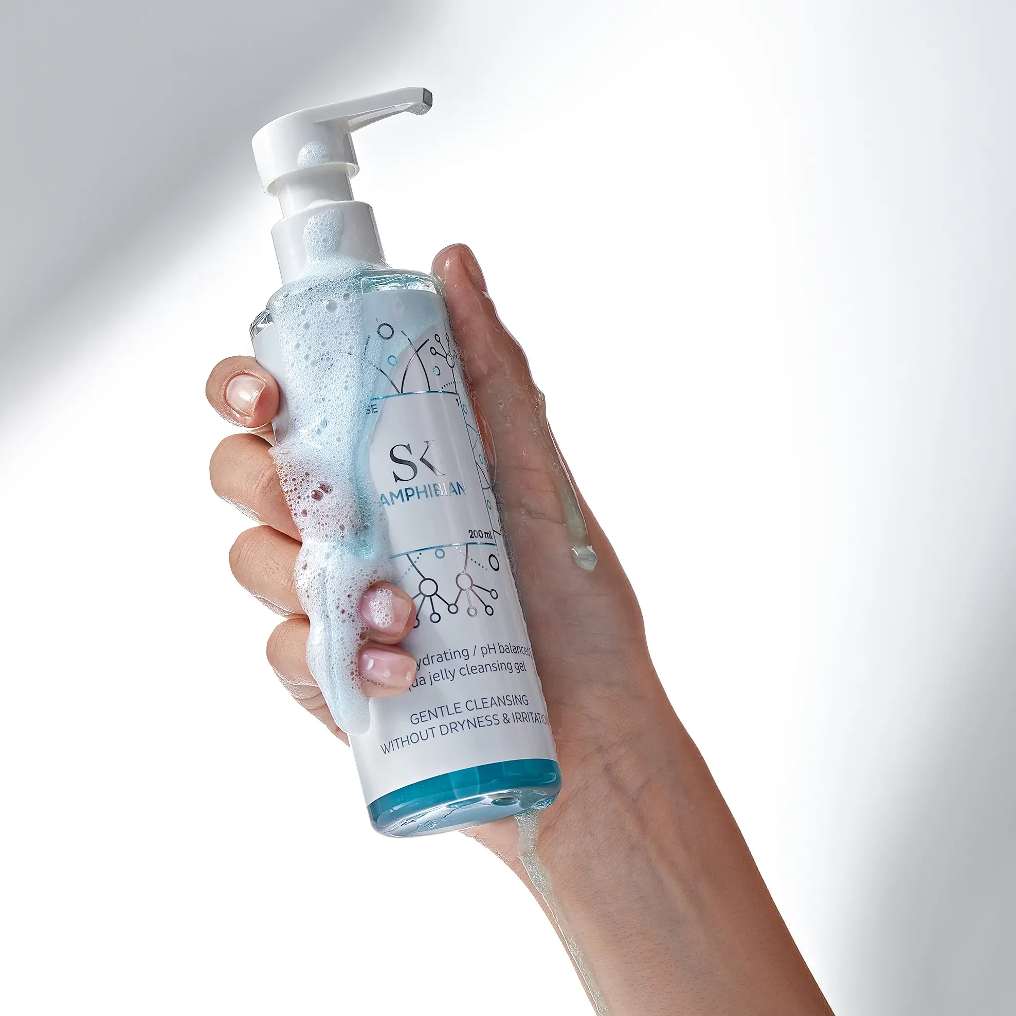 Hand holding a clear plastic bottle of hydrating face cleanser with foam spilling out, against a plain white background.