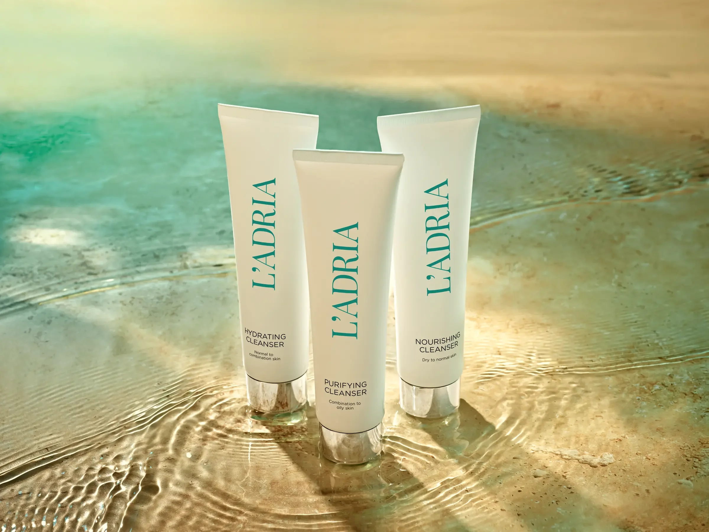 Three tubes of L'ADRIERA skincare products placed on sandy beach with gentle waves and sunlight reflection.