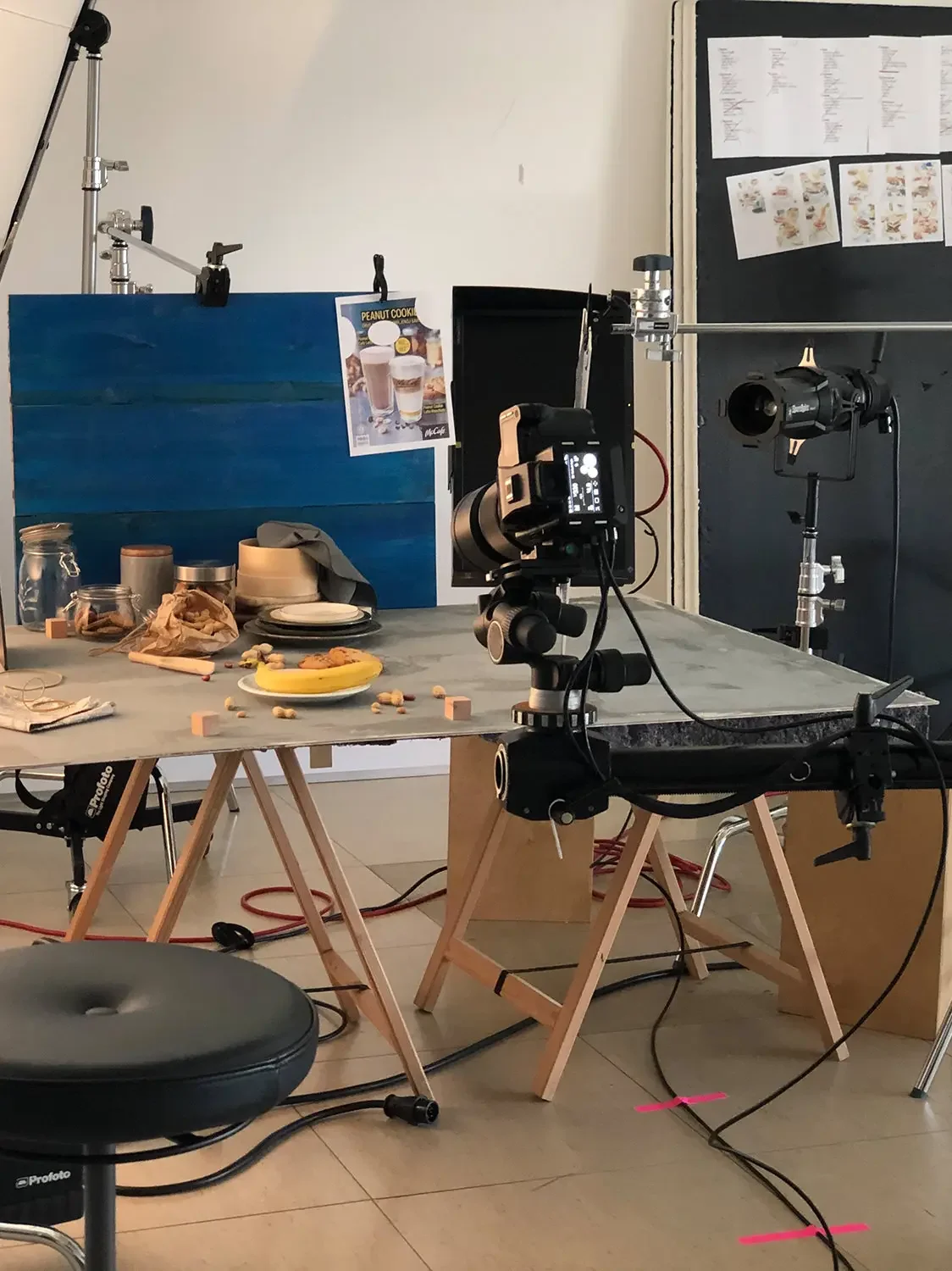 Photography setup with camera and lighting equipment on a work table, with food items, dishes, and a food poster on the wall.