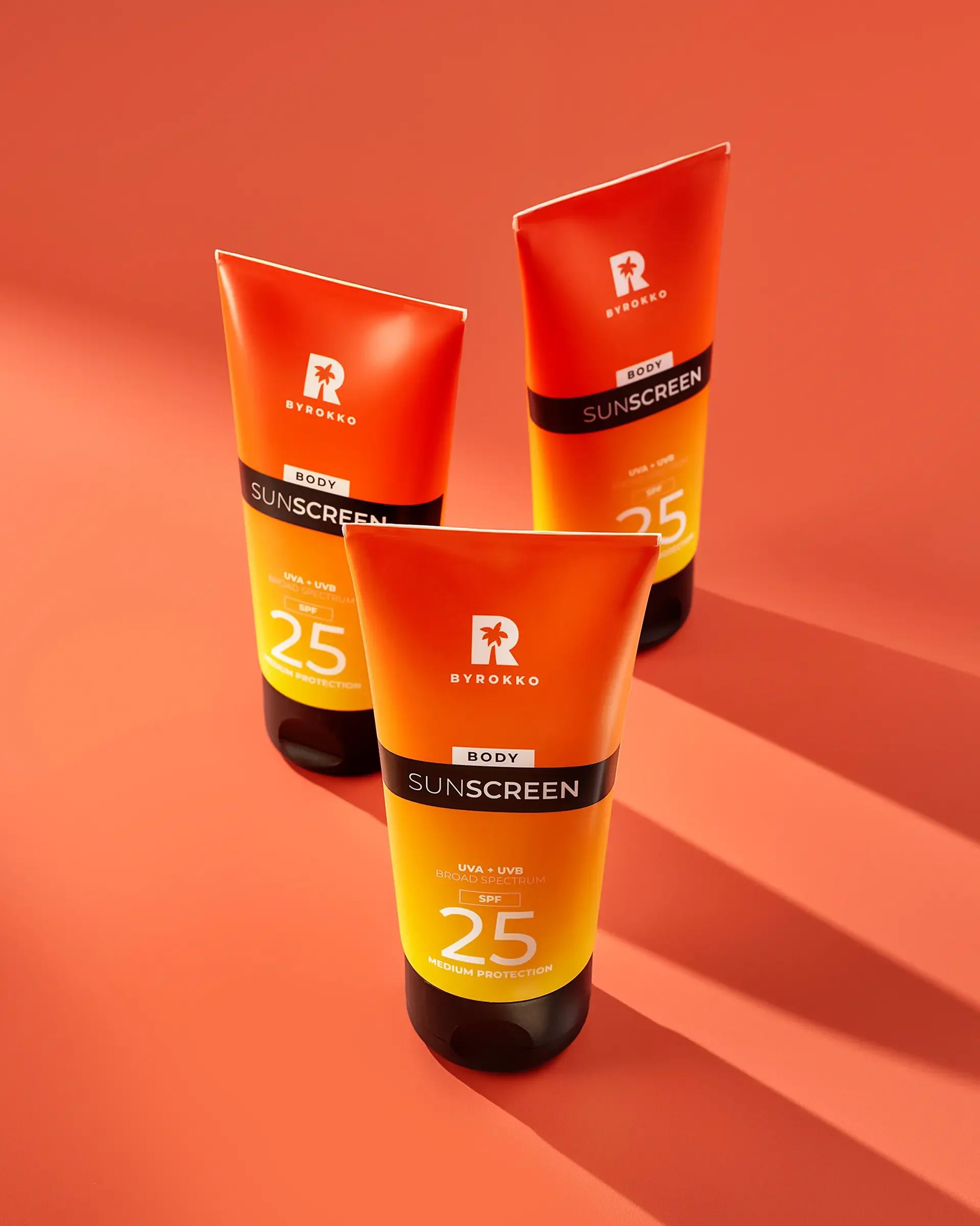 Three tubes of BYROKKO brand sunscreen standing on a pink-orange surface with shadows cast to the right.