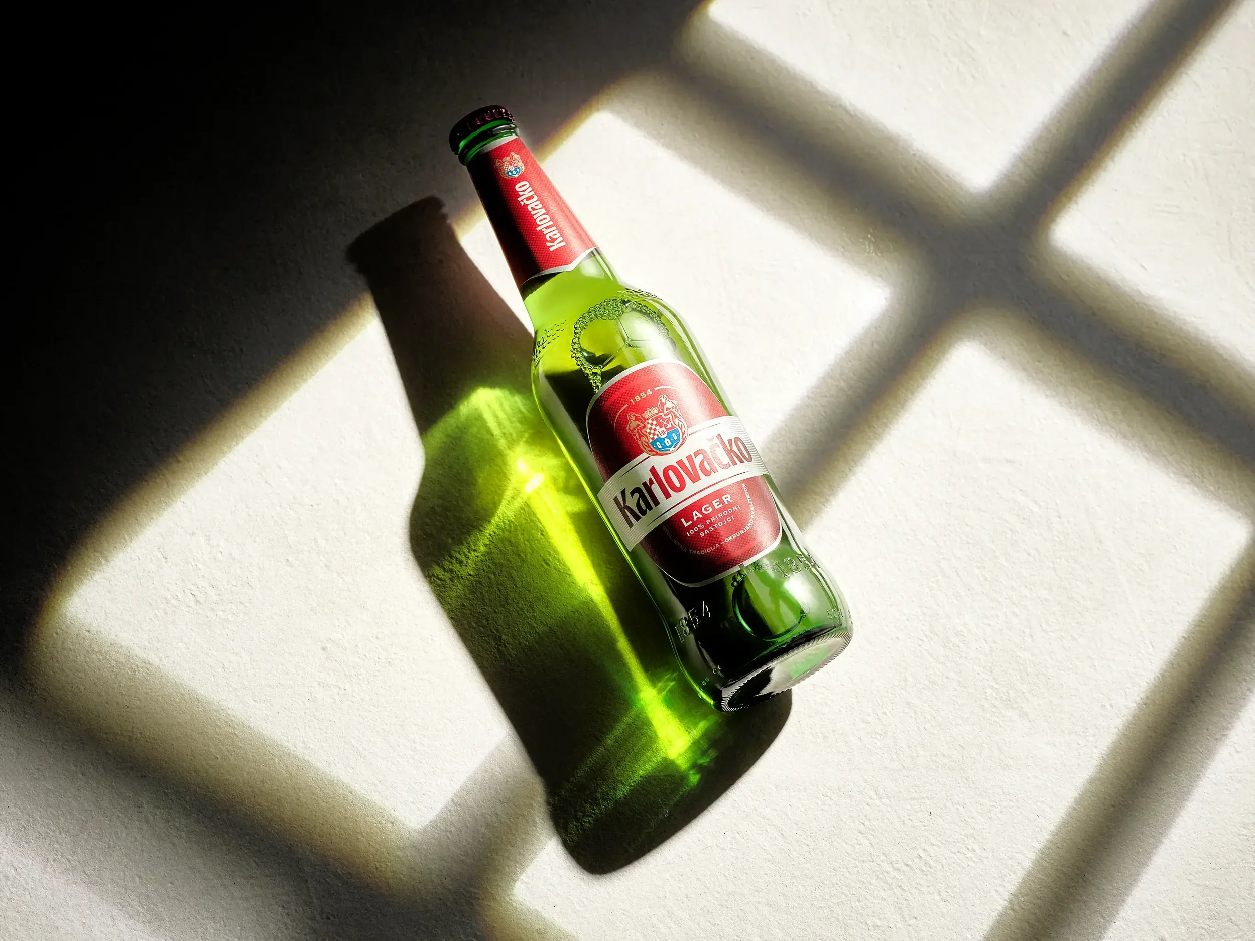 A green glass bottle of Karlovacko Lager beer lying on a white textured surface, illuminated by sunlight creating a shadow and reflection.