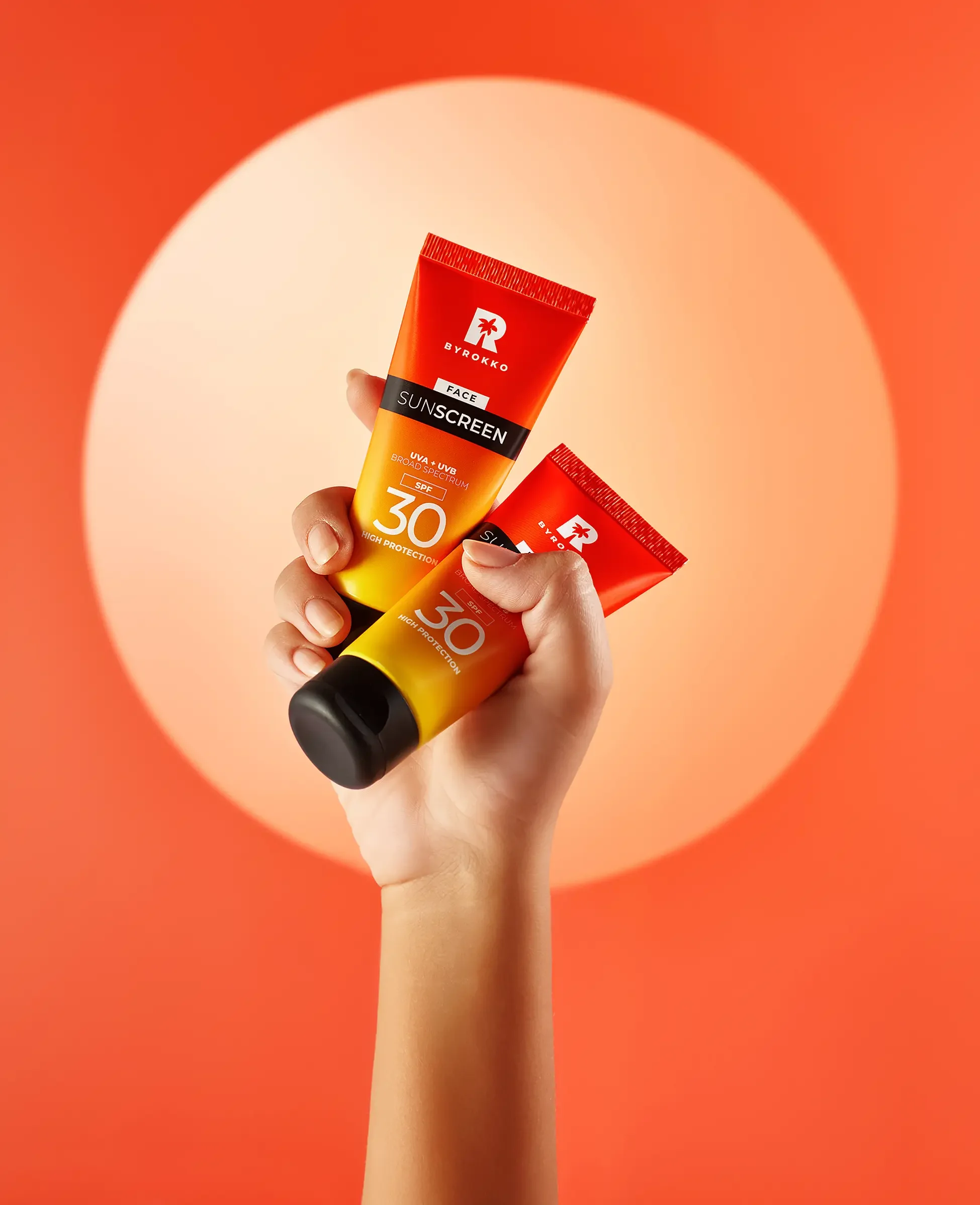 A hand holding two tubes of BYROKKO sunscreen with SPF 30 against an orange background.