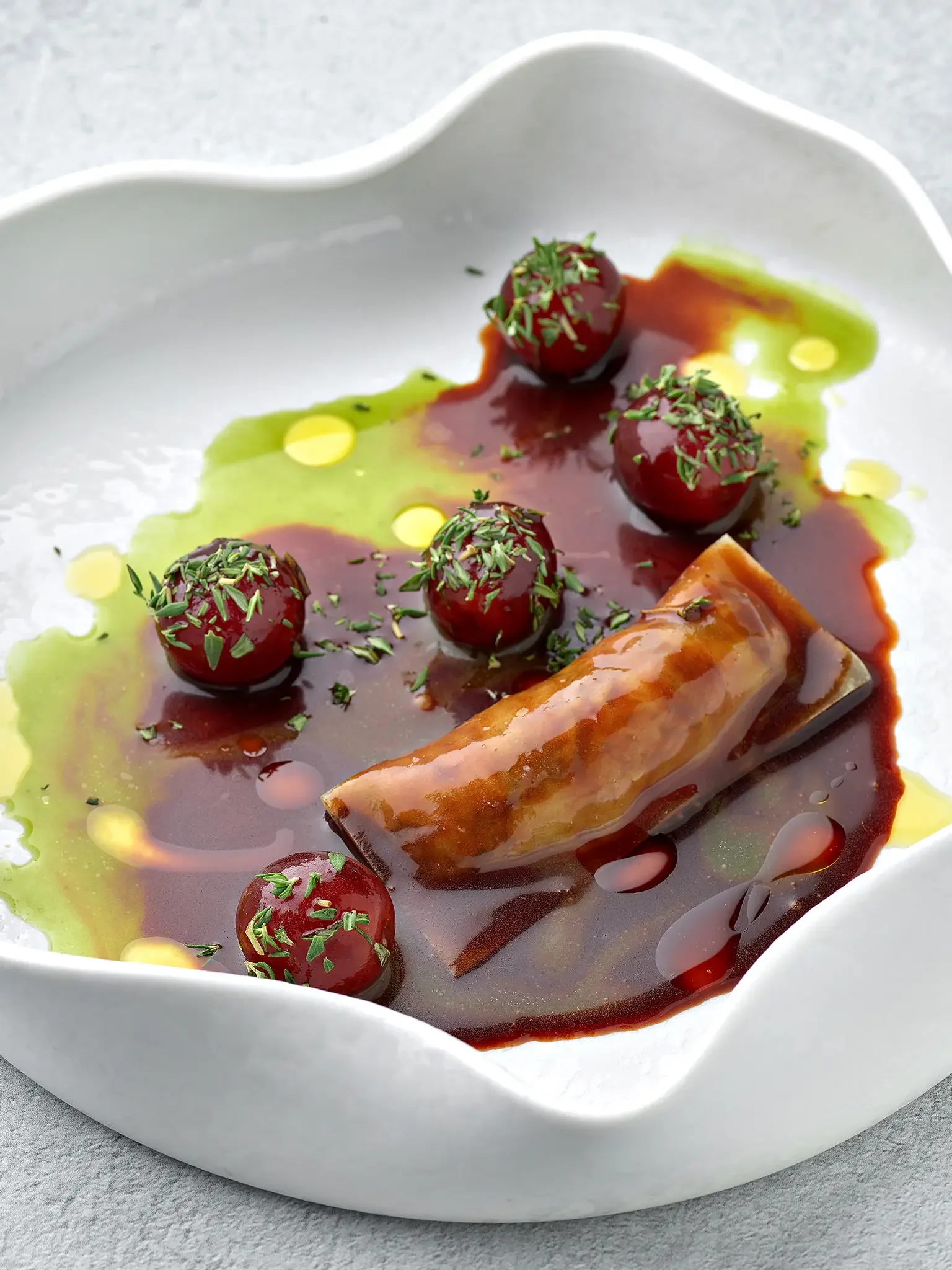 Gourmet dish in a white bowl with cherry tomatoes garnished with herbs, a piece of glazed meat or fish, and colorful sauces and oils.
