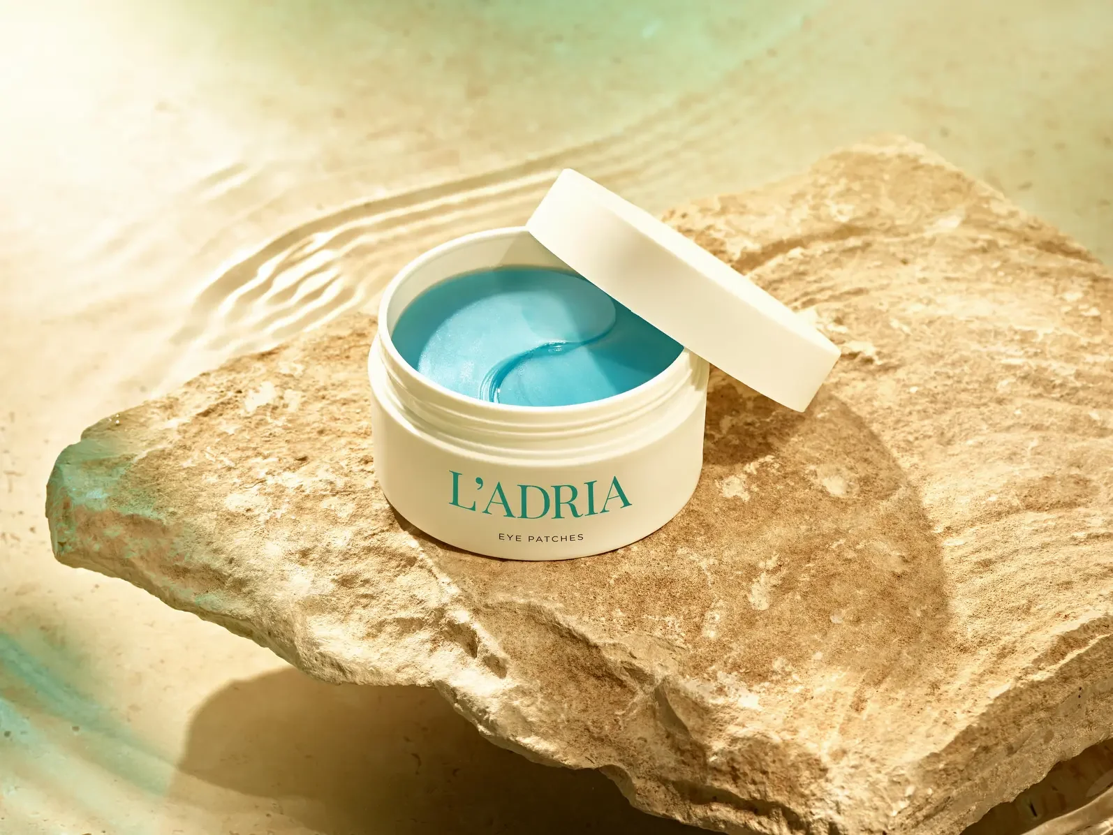 Open container of blue eye patches labeled 'L'ADRIA' on a rocky surface with a sandy background.