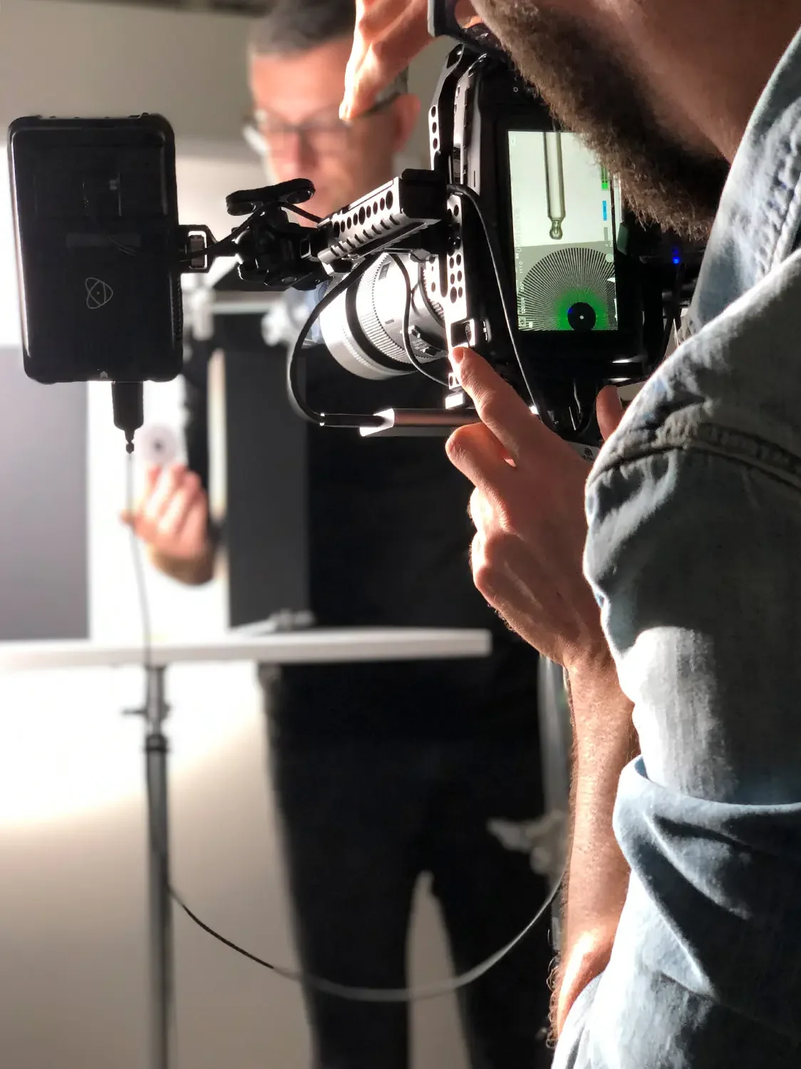 A person operating a camera on a tripod, focusing on a person in the background using a pointer or stylus in an indoor setting.