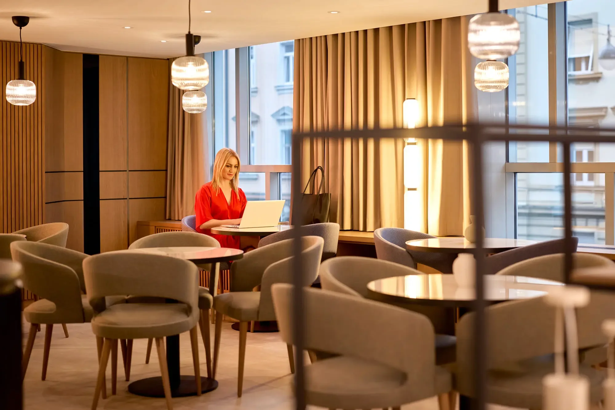 A woman sitting at a round table in a well-lit, modern café or restaurant, working on a laptop. The interior features wooden walls, beige curtains, and hanging pendant lights.