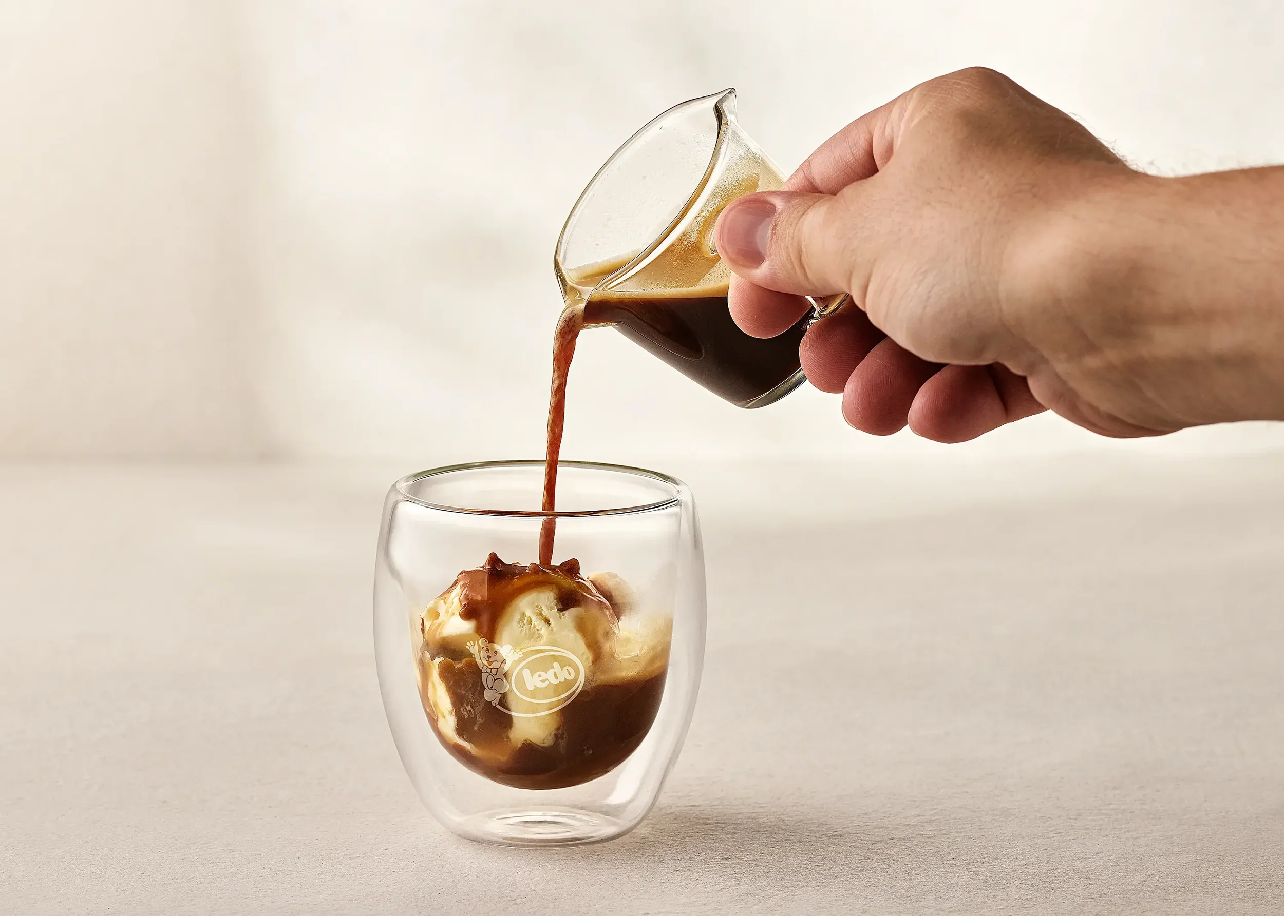 Someone pouring coffee from a glass pitcher into a clear glass cup with ice cream and chocolate sauce.