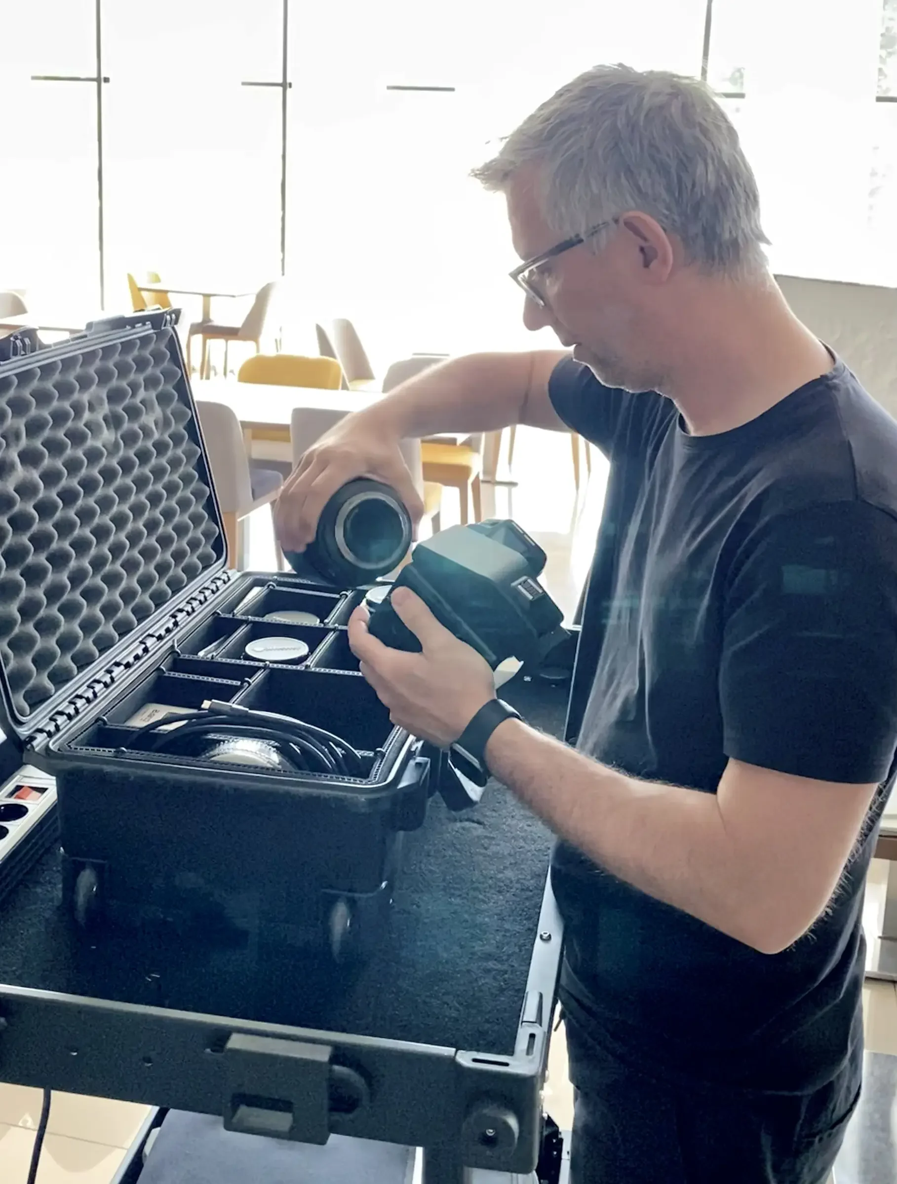 Man organizing camera equipment in a black case with foam padding inside a well-lit room with large windows and chairs in the background.