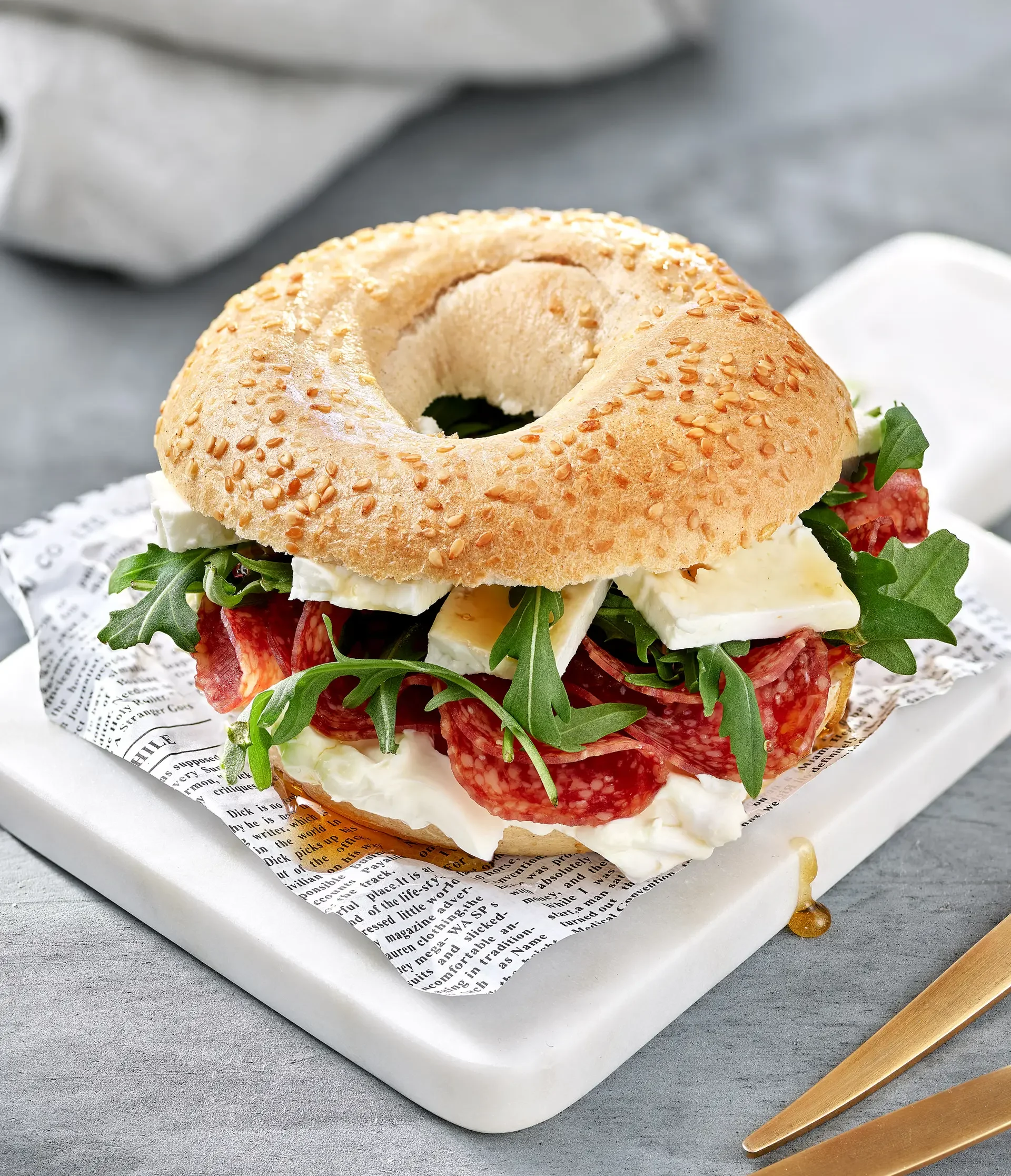 Bagel sandwich with salami, arugula, cheese, and cream cheese on a tray with newspaper-like paper and a gold fork.