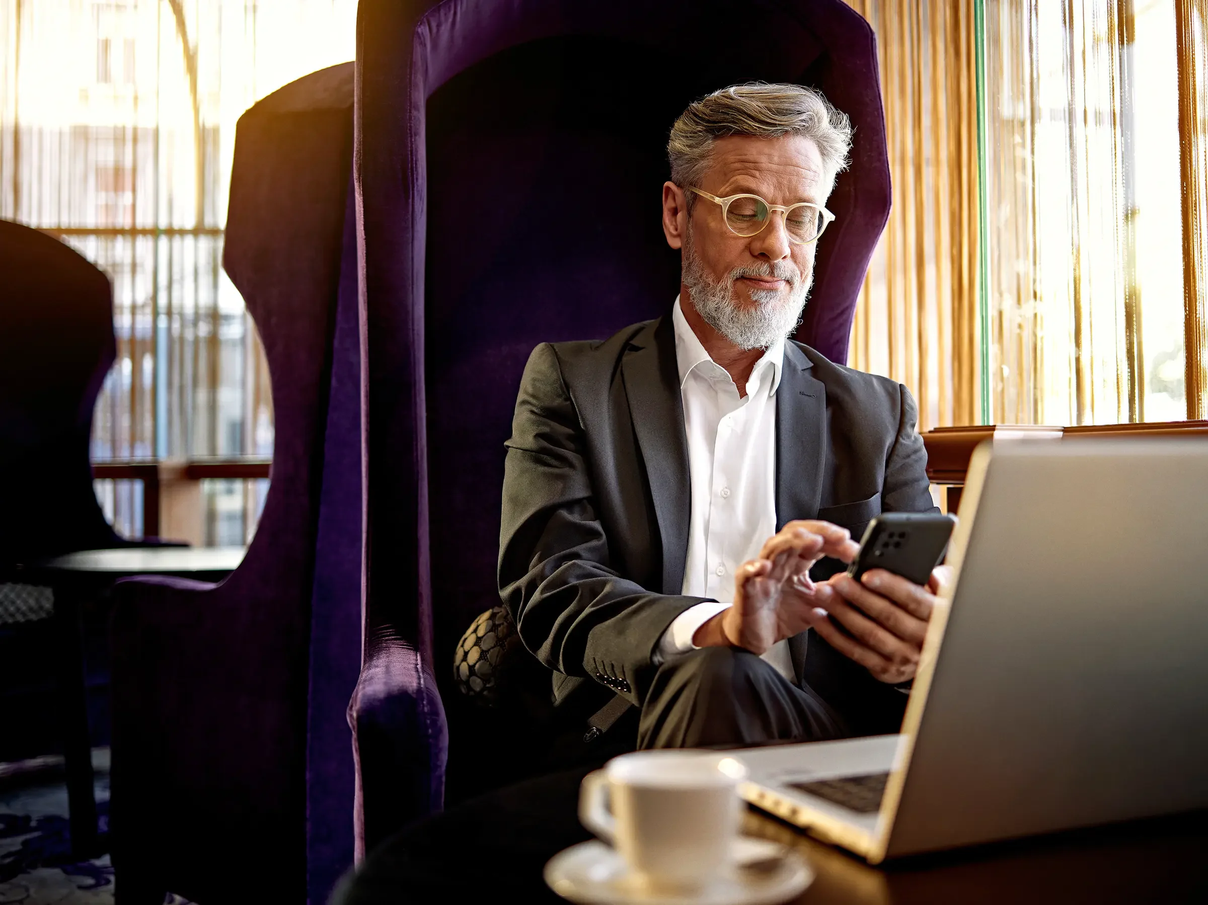 An older man with gray hair and beard wearing glasses and a business suit sitting in a high-backed purple chair, looking at his smartphone, with a laptop and coffee cup on the table in front of him, in a sunlit room with bamboo blinds.