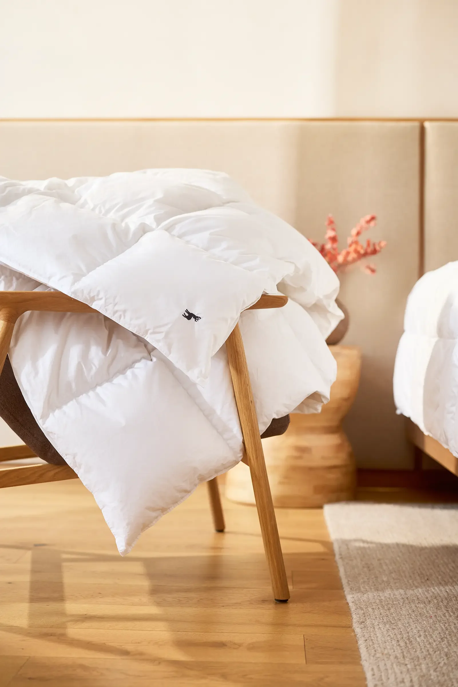 A cozy bed with white bedding and a pillow on a wooden bed frame in a bedroom with a wooden nightstand and a pink flower arrangement in a woven basket.