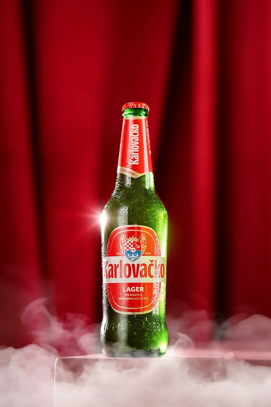 A green bottle of Karlovacko Lager beer with condensation, placed on a clear stand with fog or dry ice around the base, against a red curtain background.