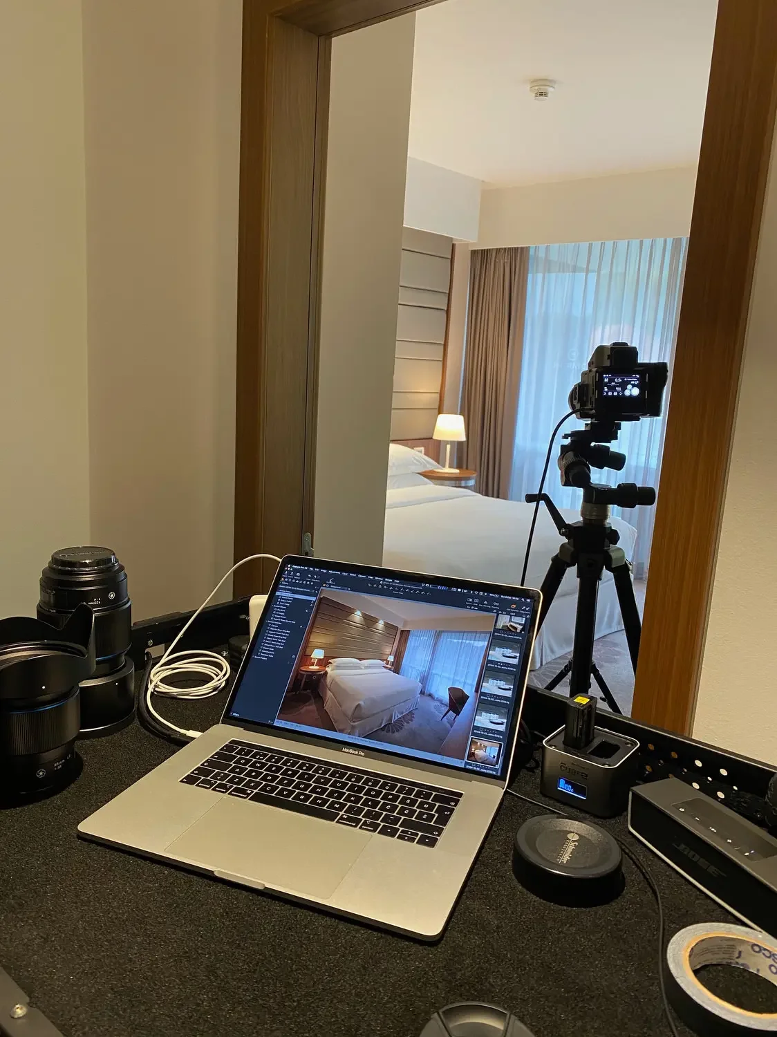 A hotel room is being photographed, with a camera mounted on a tripod. The view is from a workspace with a laptop displaying the room's image, a camera lens, and various photography accessories on a black table.