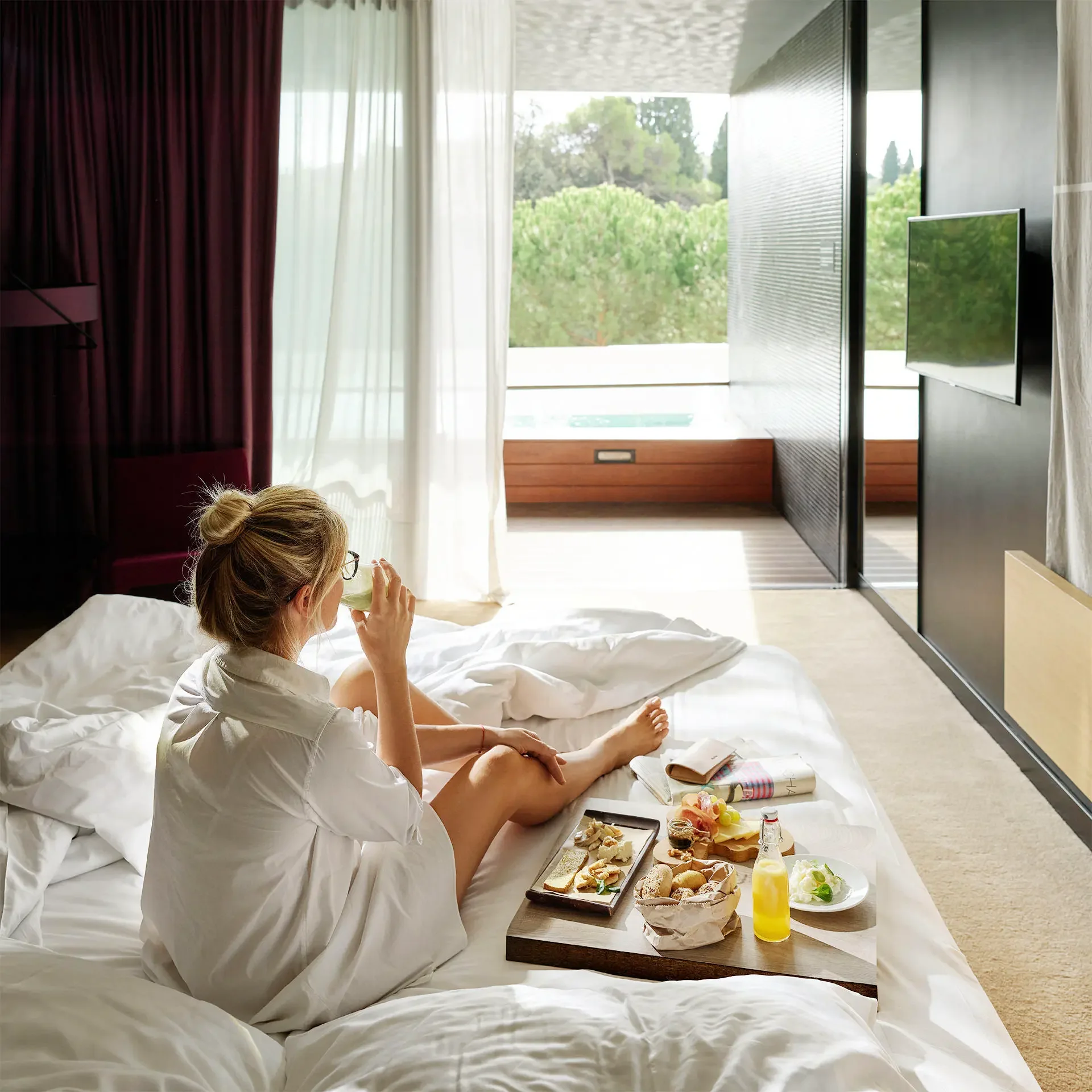 A woman sitting on a bed having breakfast with bread, pastries, cheese, juice, and a salad in a modern hotel room with large windows and a view of greenery outside.