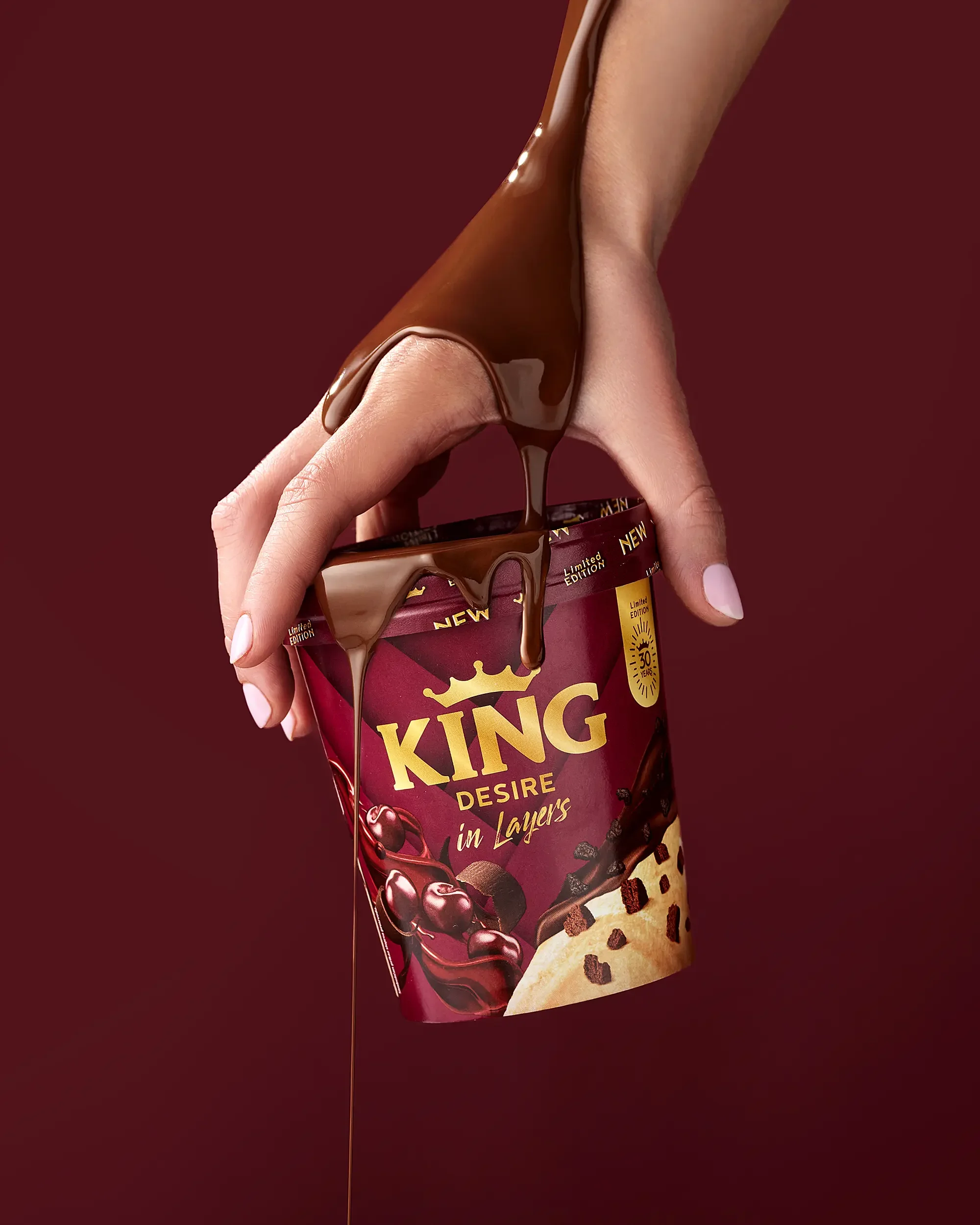 Hand holding a cup of King Desire ice cream with chocolate sauce pouring over it, dark red background.