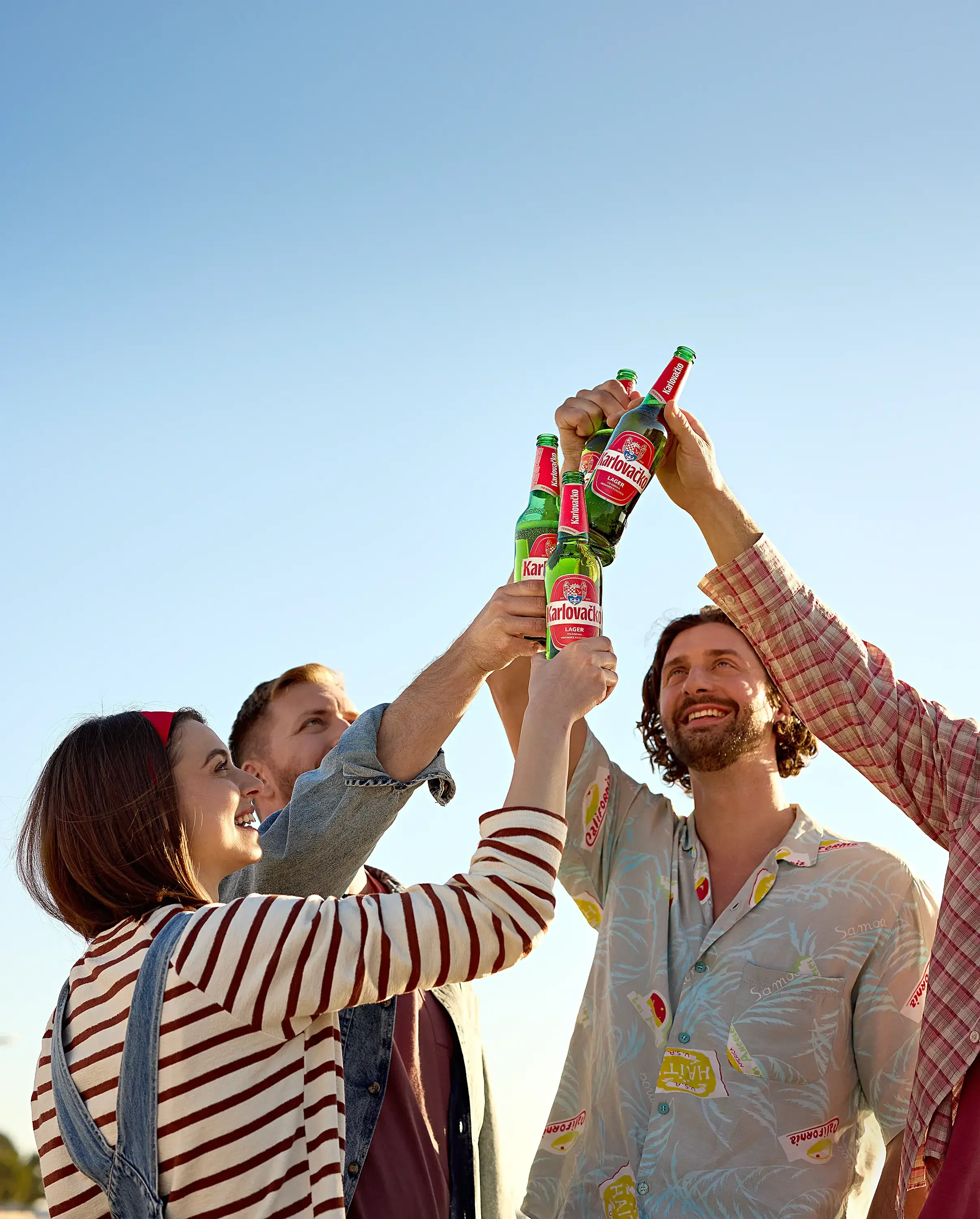 Group of four friends raising bottles of Karlovačko beer in a toast outdoors on a sunny day.