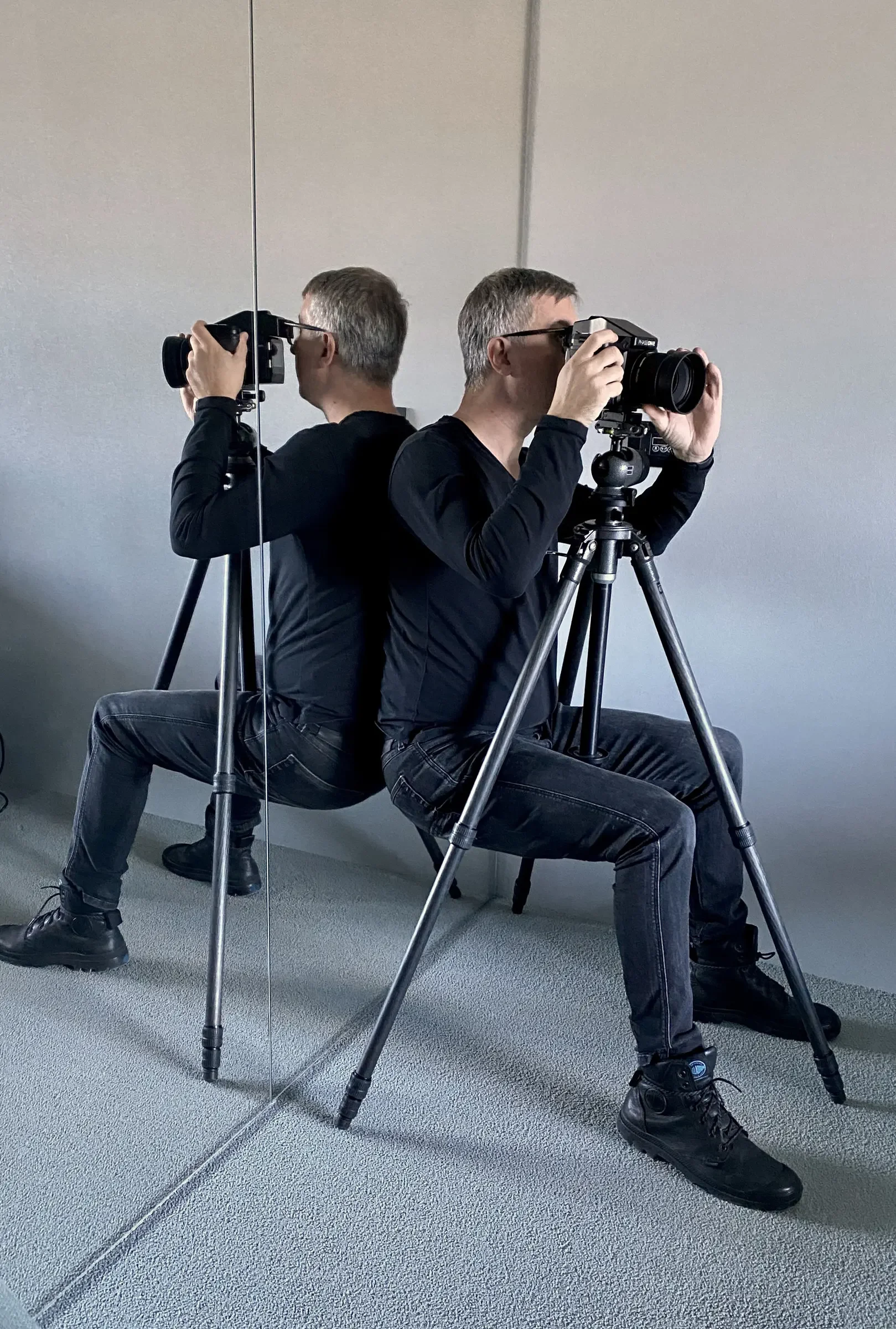 A person taking a photograph with a professional camera mounted on a tripod, sitting back-to-back against a mirror.