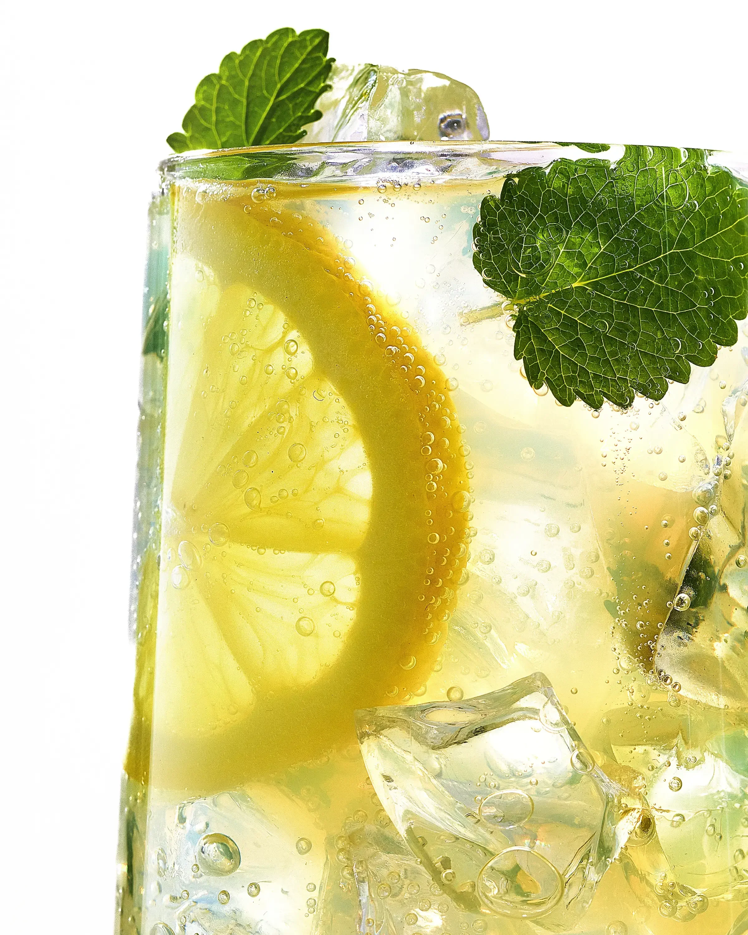 Close-up of a glass of lemon and mint flavored sparkling water with lemon slices, mint leaves, ice cubes, and bubbles.