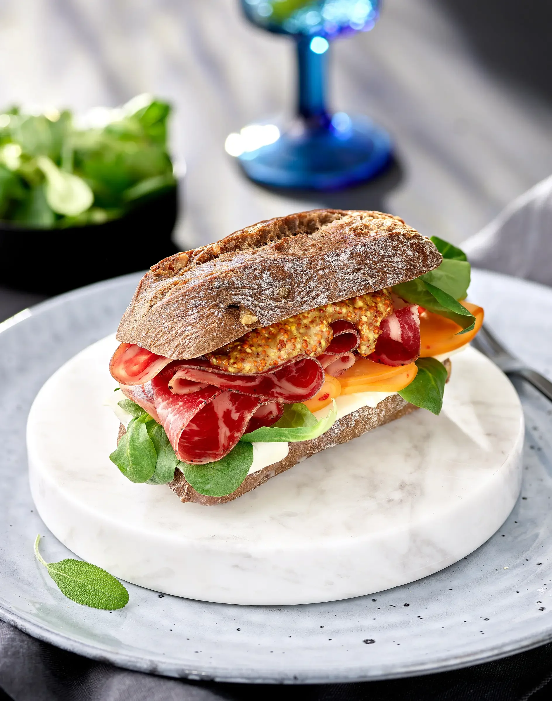 A sandwich on a round white plate, with leafy greens, sliced tomatoes, various deli meats, mustard, and a rustic baguette.