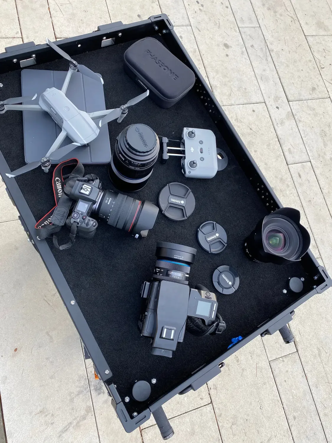 A black carry case with various professional camera equipment, including a drone, a DSLR camera, a lens, a gimbal, and multiple camera lenses, all arranged on a black surface in an outdoor setting.