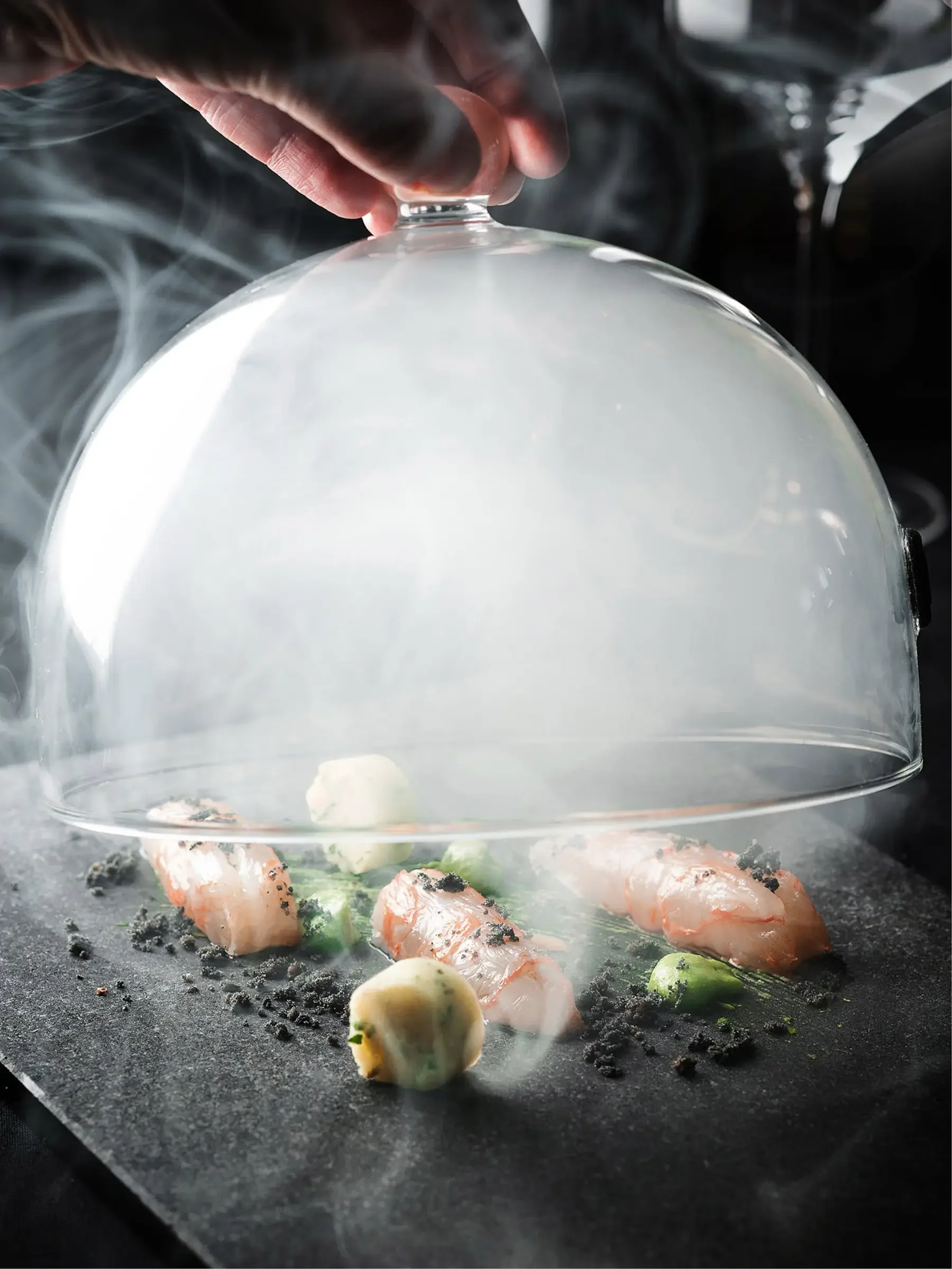 Chef lifting glass dome off of a dish with shrimp, garlic, and green peas, garnished with black crumbs, on a black surface with steam and smoke.