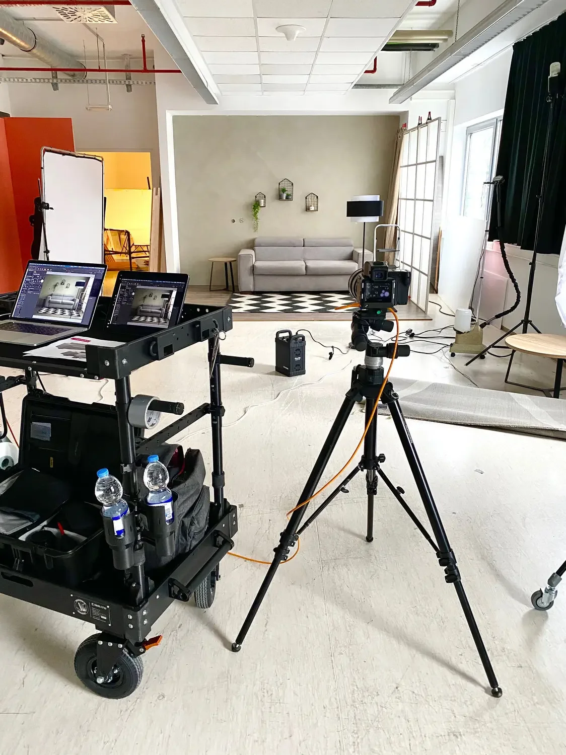 Photography studio setup with a sofa, lighting, camera, and two monitors, in front of a painted wall with houseplant decorations.