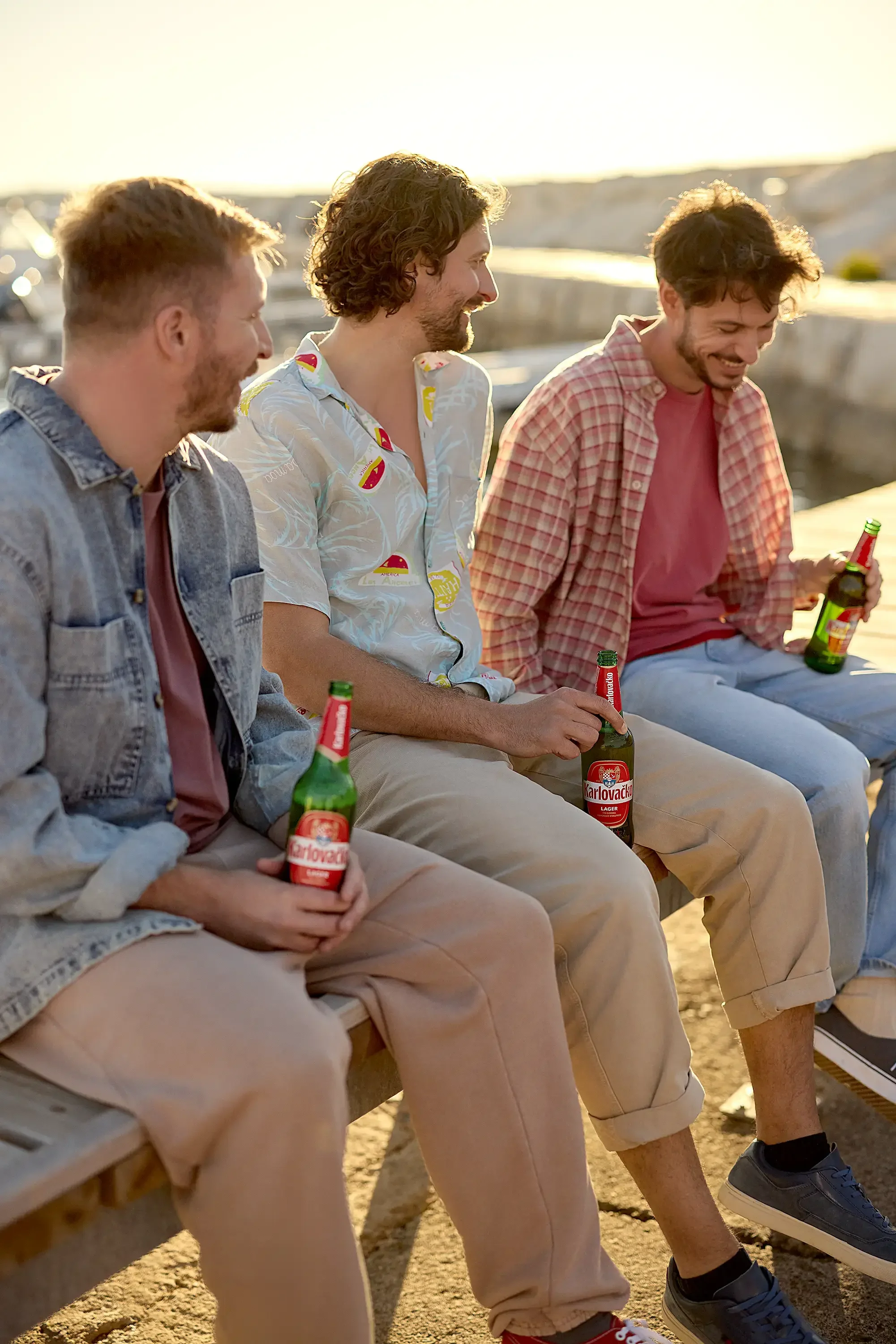 Three young men sitting on a bench by the water, holding bottles of Karlovacko beer, enjoying sunset, smiling, dressed casually, with a marina and boats in the background.