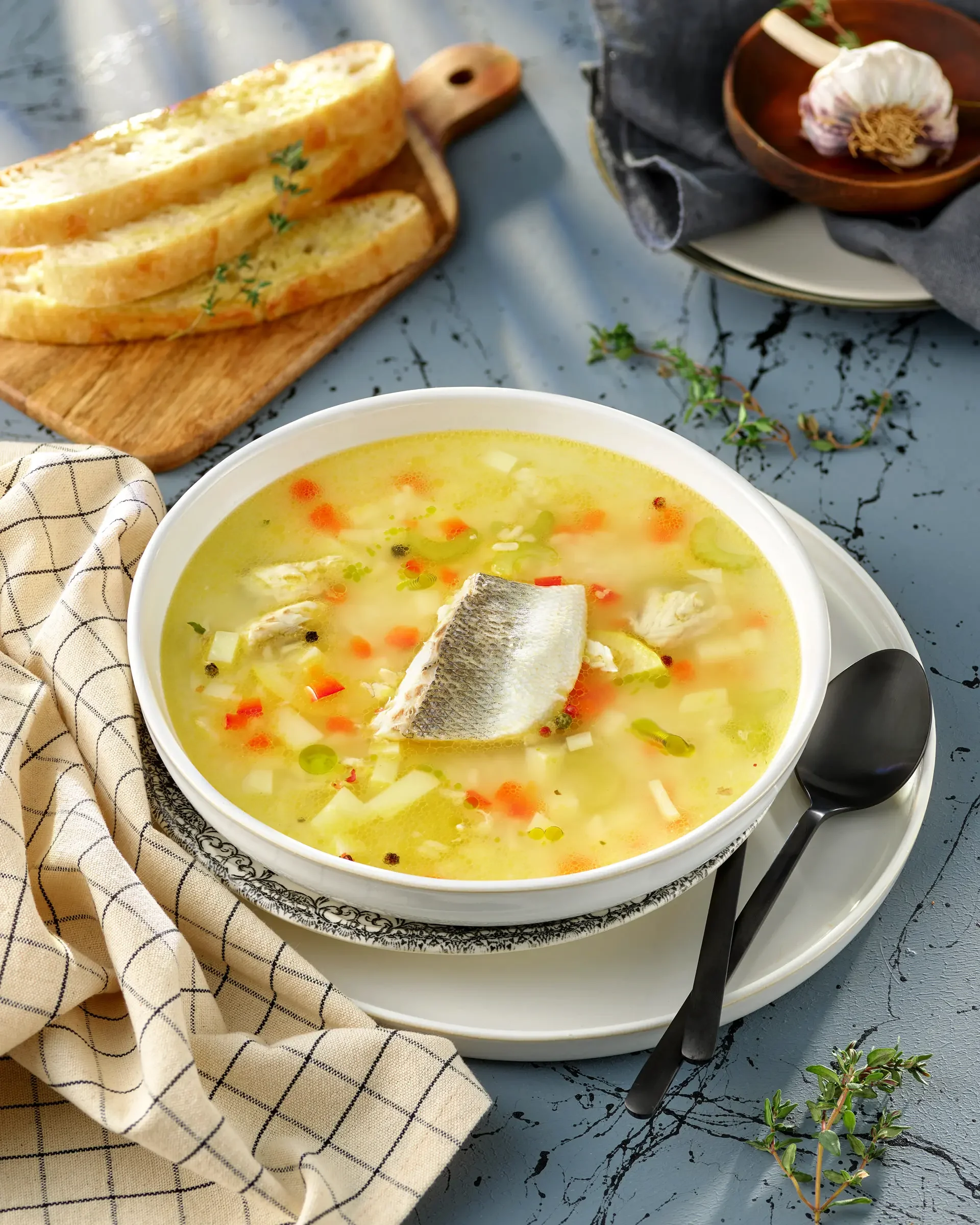 A bowl of fish soup with a piece of fish on top, served with slices of bread on a wooden cutting board nearby.