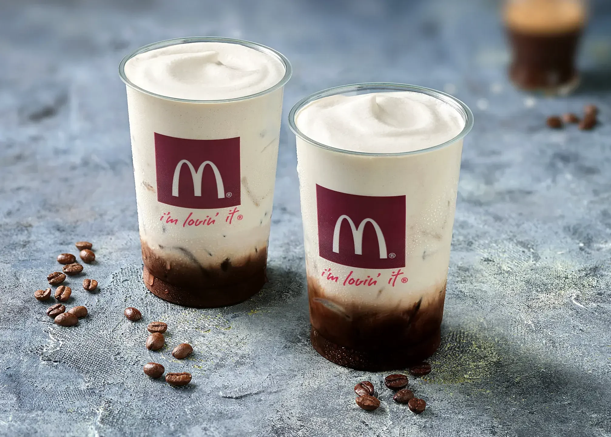 Two McDonald's frappé drinks with whipped cream on top, surrounded by coffee beans on a textured gray surface.