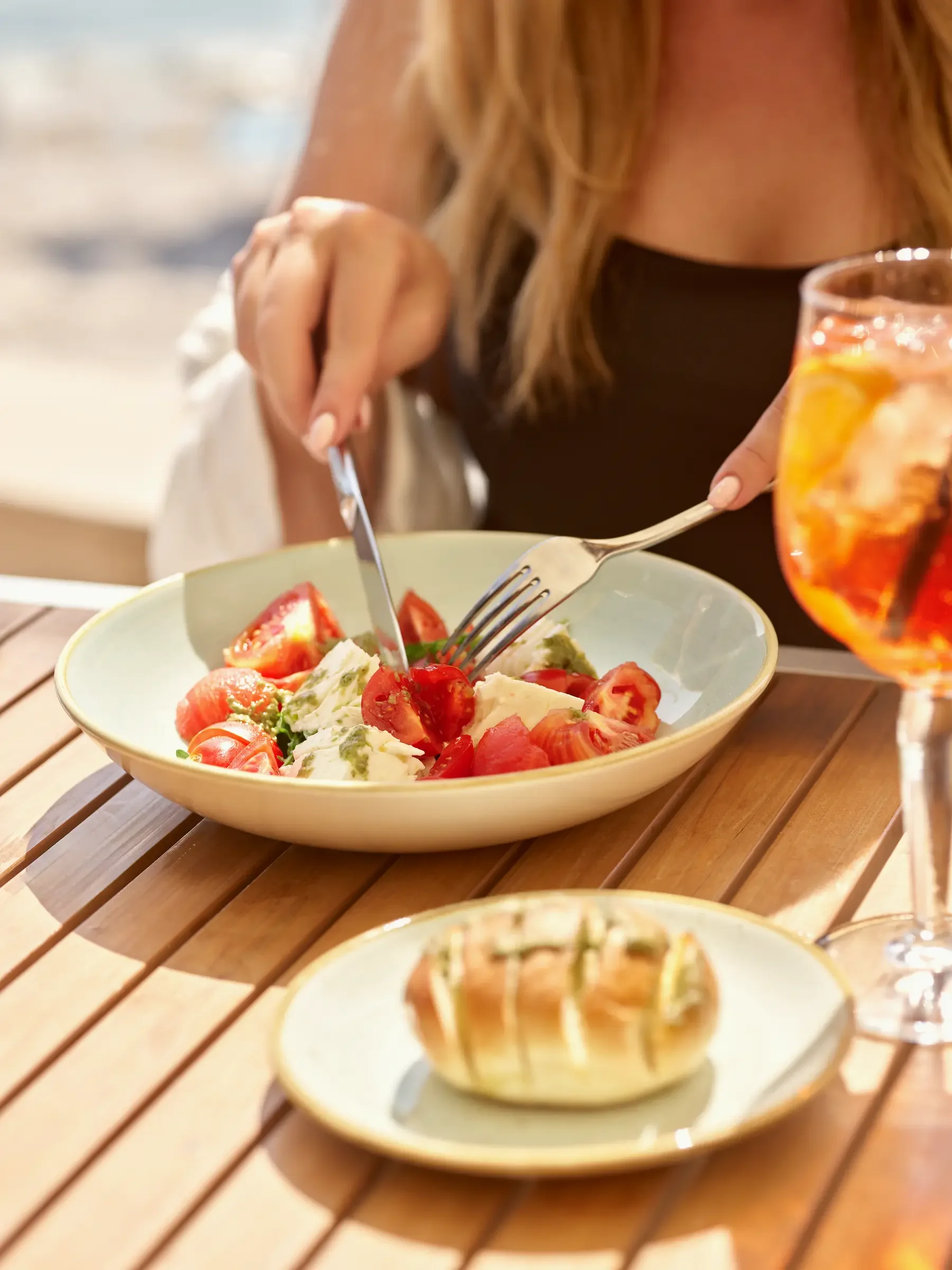 A woman eating a fresh salad with tomatoes, mozzarella, and herbs at an outdoor table, with a glass of iced drink nearby.