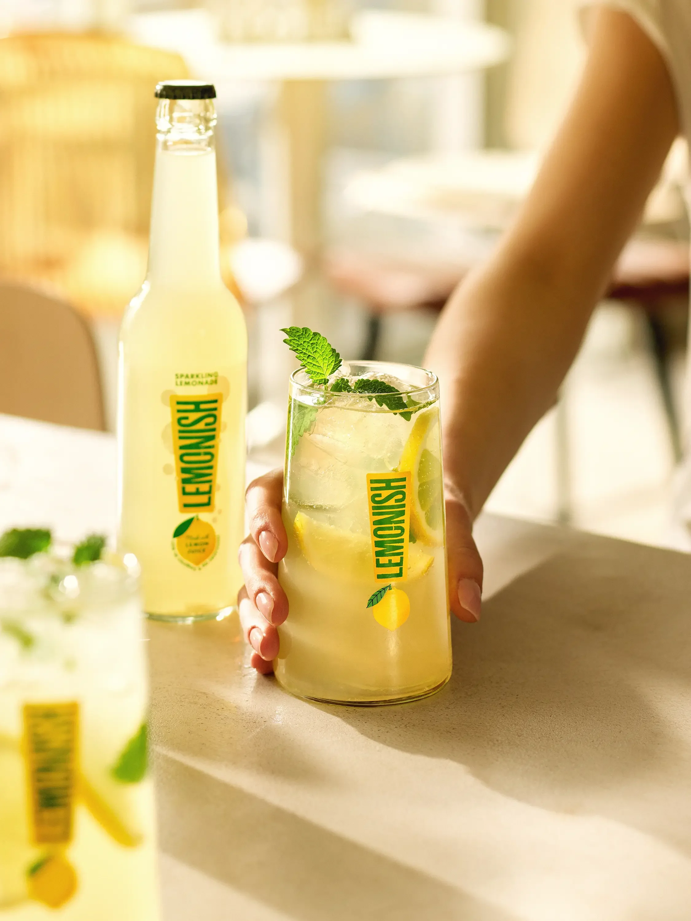 A person holding a glass of lemonade with ice, lemon slices, and mint leaves. There is a lemon-flavored drink bottle in the background.