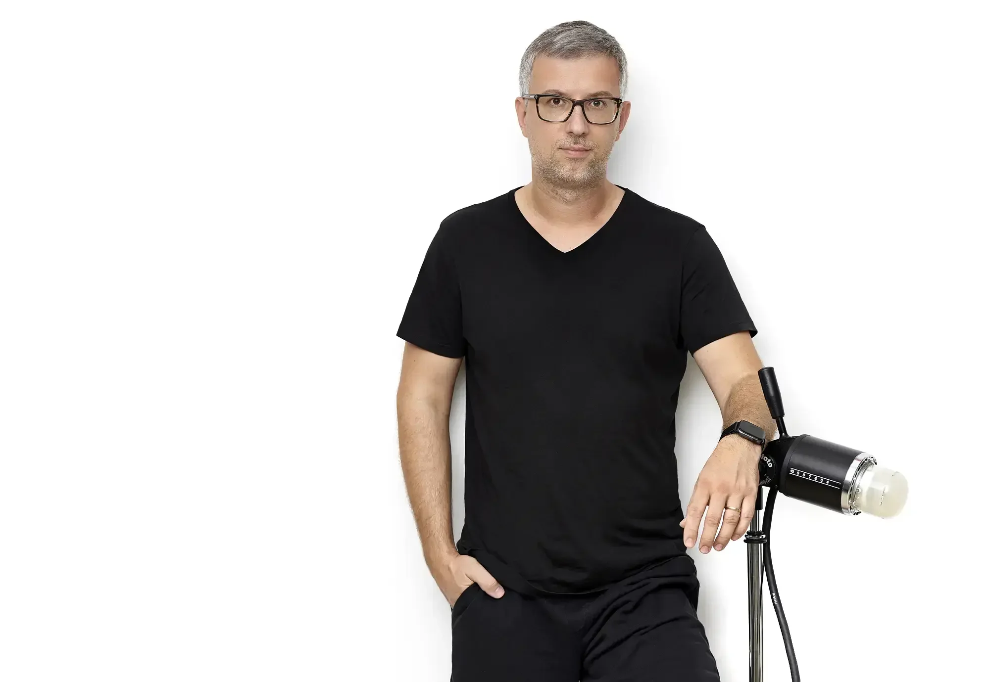 A man with short gray hair, glasses, and a beard wearing a black t-shirt and black pants, standing against a white background with a studio light on a stand.