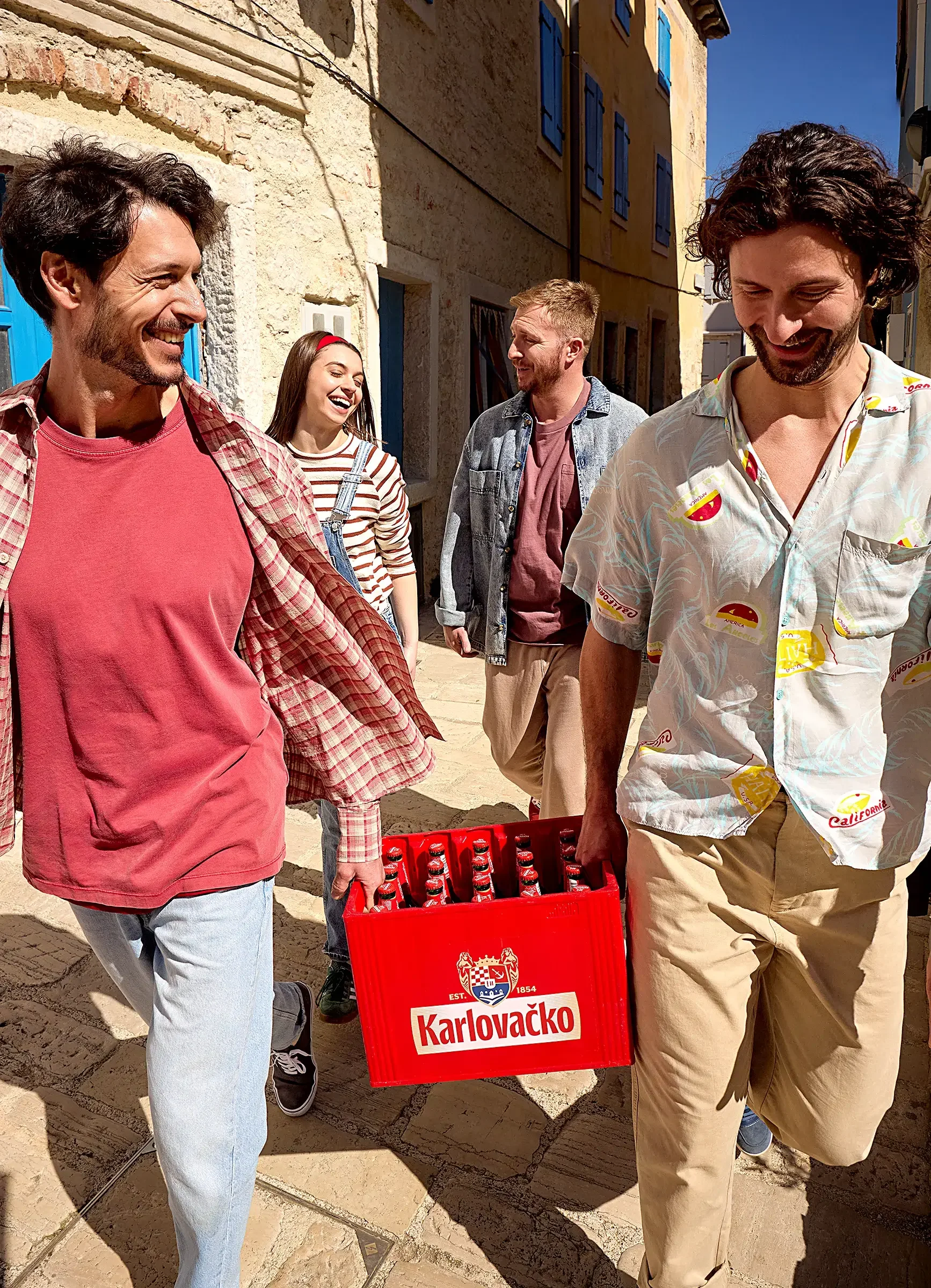A group of four friends walking down a sunny street, carrying a crate of Karlovačko beer and smiling.