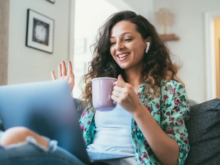 A BIPOC female coach seated with legs up on a grey charcoal coach with a laptop.