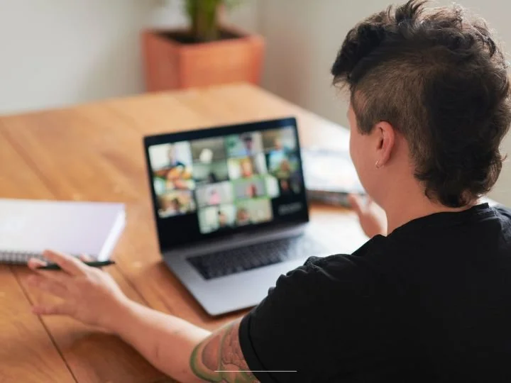 A coach or therapist with a mohawk with their back towards the photographer facing a laptop screen of a group of people while seated at their wooden table with a pad of paper and pen.