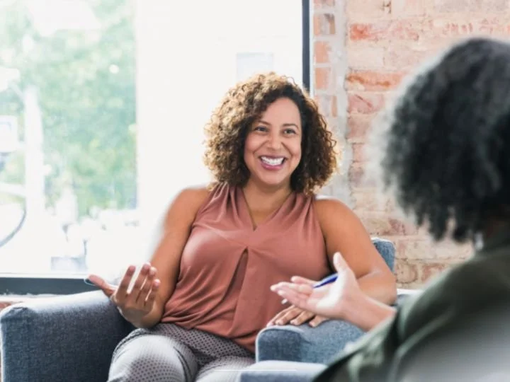 Two adult Black women seated in charcoal grey upholstered chairs  having a coaching conversation in a light filled room with exposed brick.