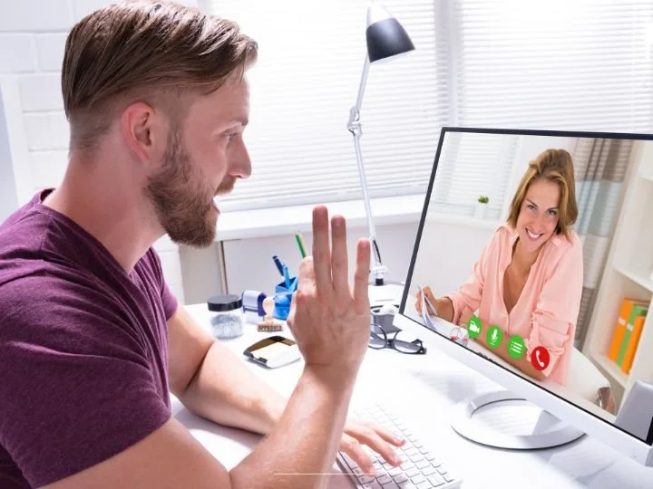 A white bodied female Life Coach joins a virtual coaching session in a beach blouse at her desk in a room with light and windows with long curtains. Her client is a white bodied male in the foreground of the photo seated a a desk with a small lamp.