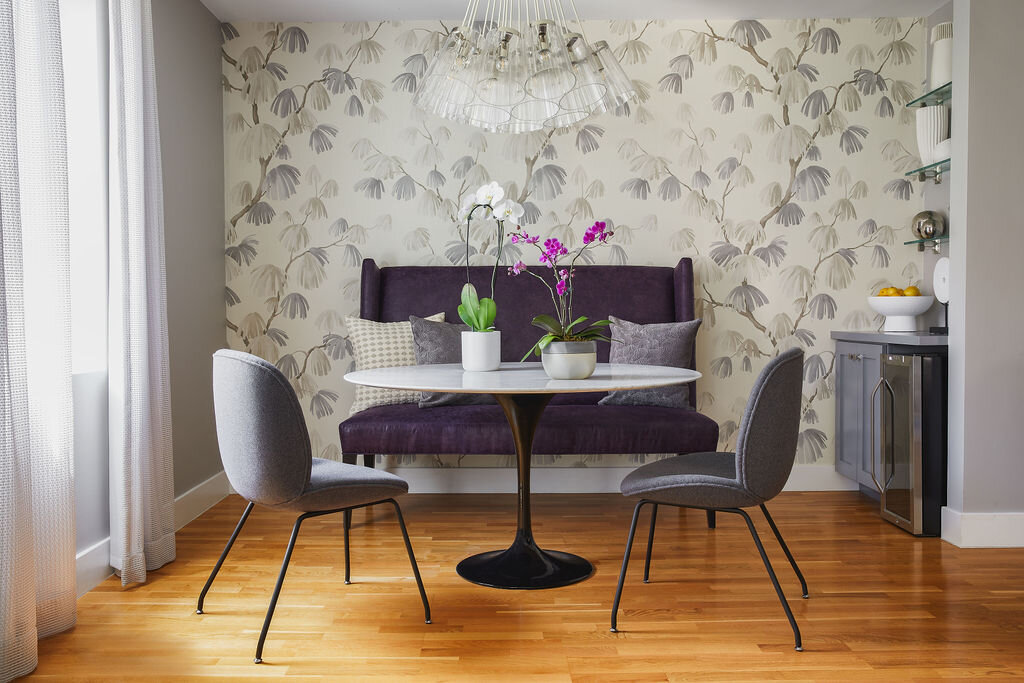 Dining area with a round white table, purple and gray chairs, a purple upholstered bench, and potted orchids, against a patterned wallpaper wall and wooden floor.