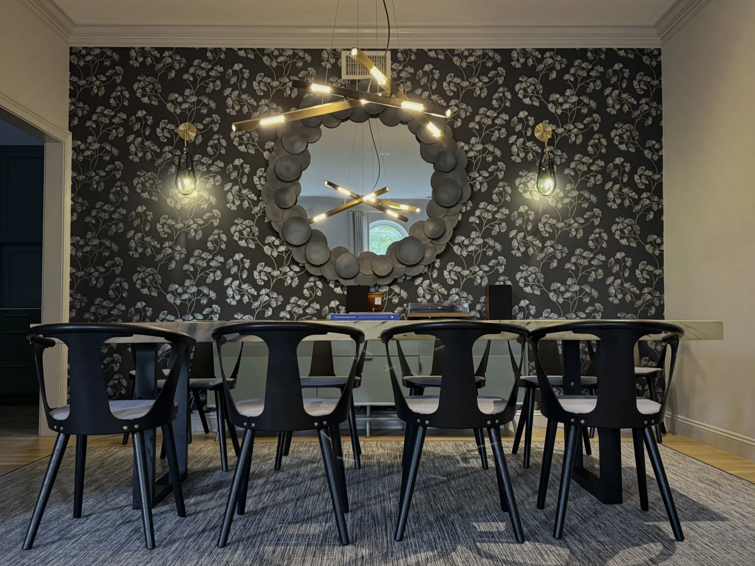 Ginkgo leaves climb a dark wallpaper, the kind of pattern that could read traditional on its own. Then the room gets modern: a live edge table, black chairs with clean lines, a chandelier of crossed mixed metal rods, sculptural and edgy where the wal