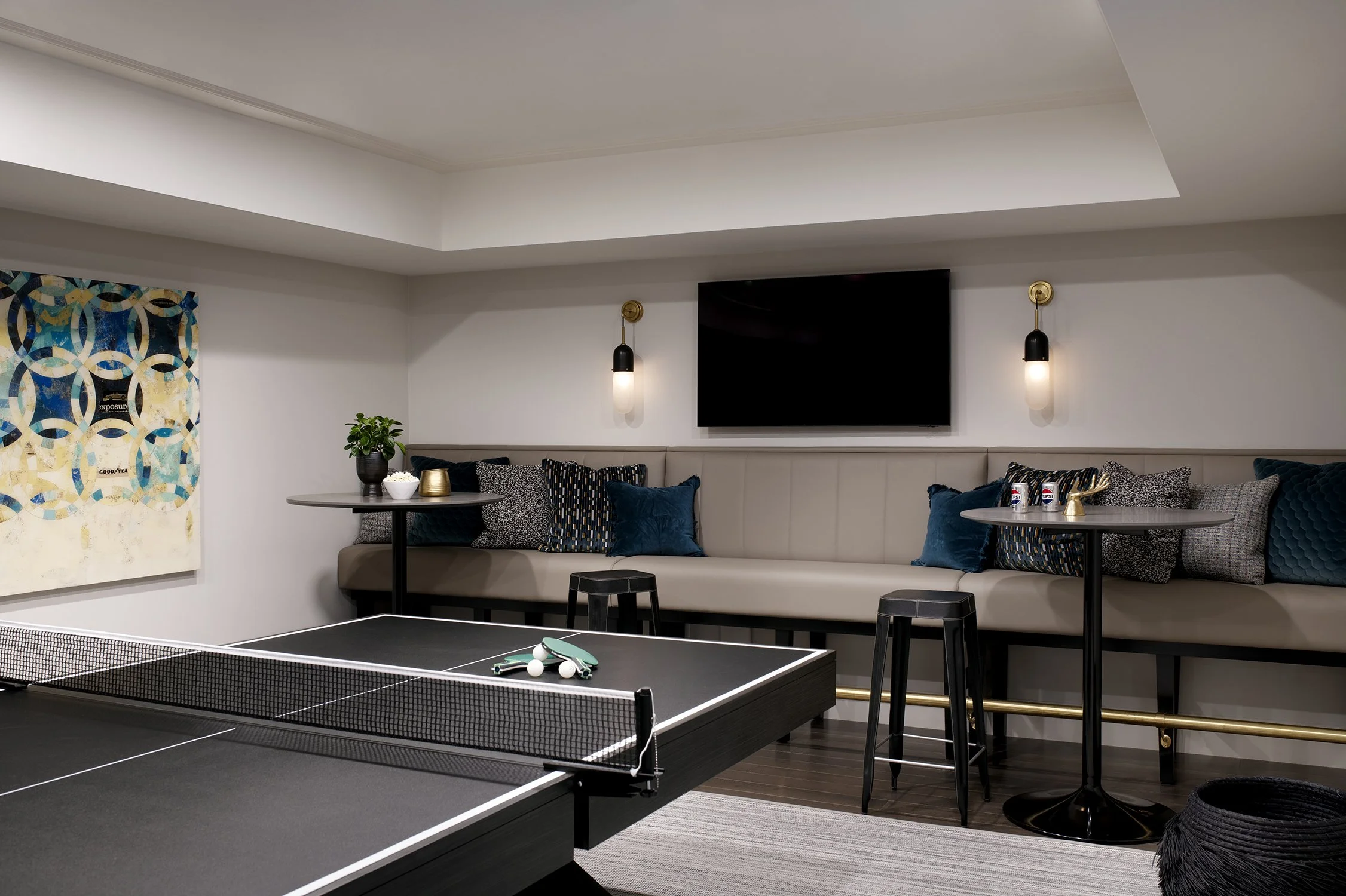 Indoor recreational room with a ping pong table, a wall-mounted TV, a long beige cushioned bench with decorative pillows, black high stools, small round tables, and a colorful wall art piece on white walls with pendant lighting.