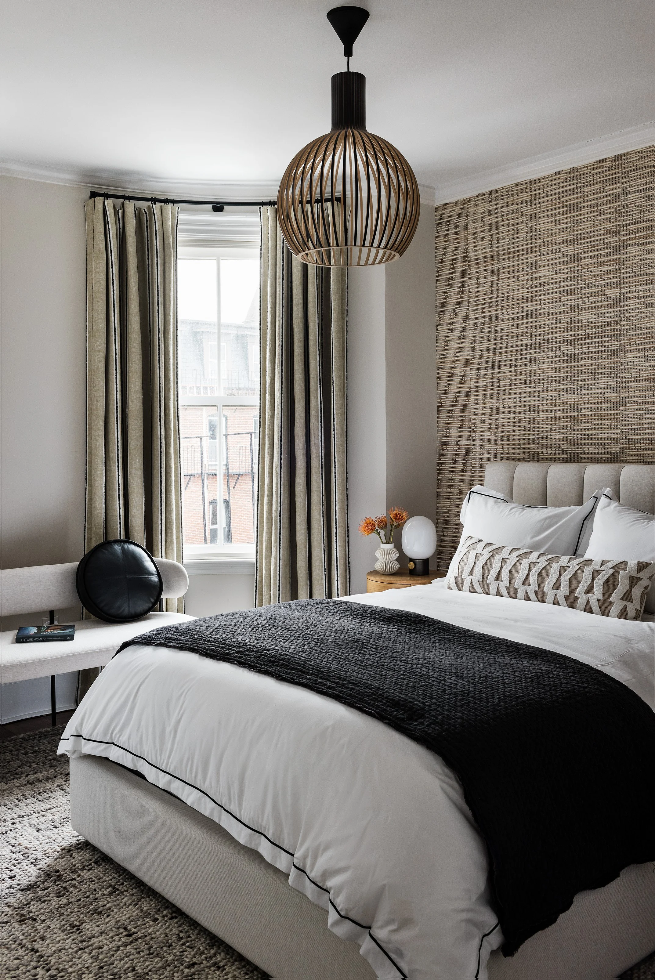 Modern bedroom with a white upholstered bed, beige headboard, and white bedding with black accents. Nightstand with a vase of orange flowers and a unique white lamp. Light-colored curtains and a large window behind. A decorative hanging pendant light