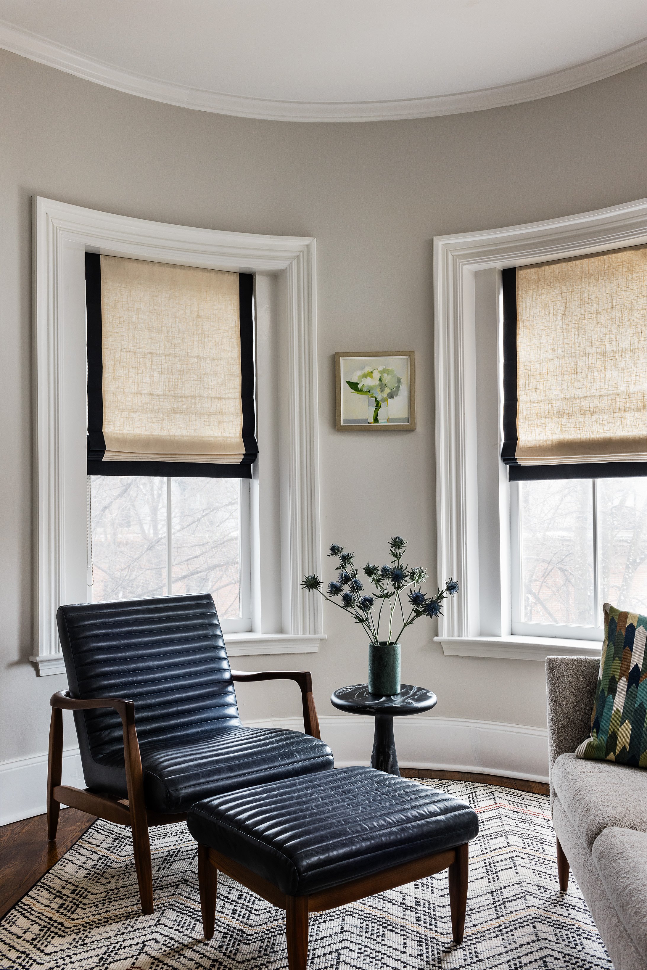A cozy living room corner with a black leather lounge chair and matching ottoman, a small black side table with a flower vase, beige window shades, a framed painting of flowers, and a patterned rug.