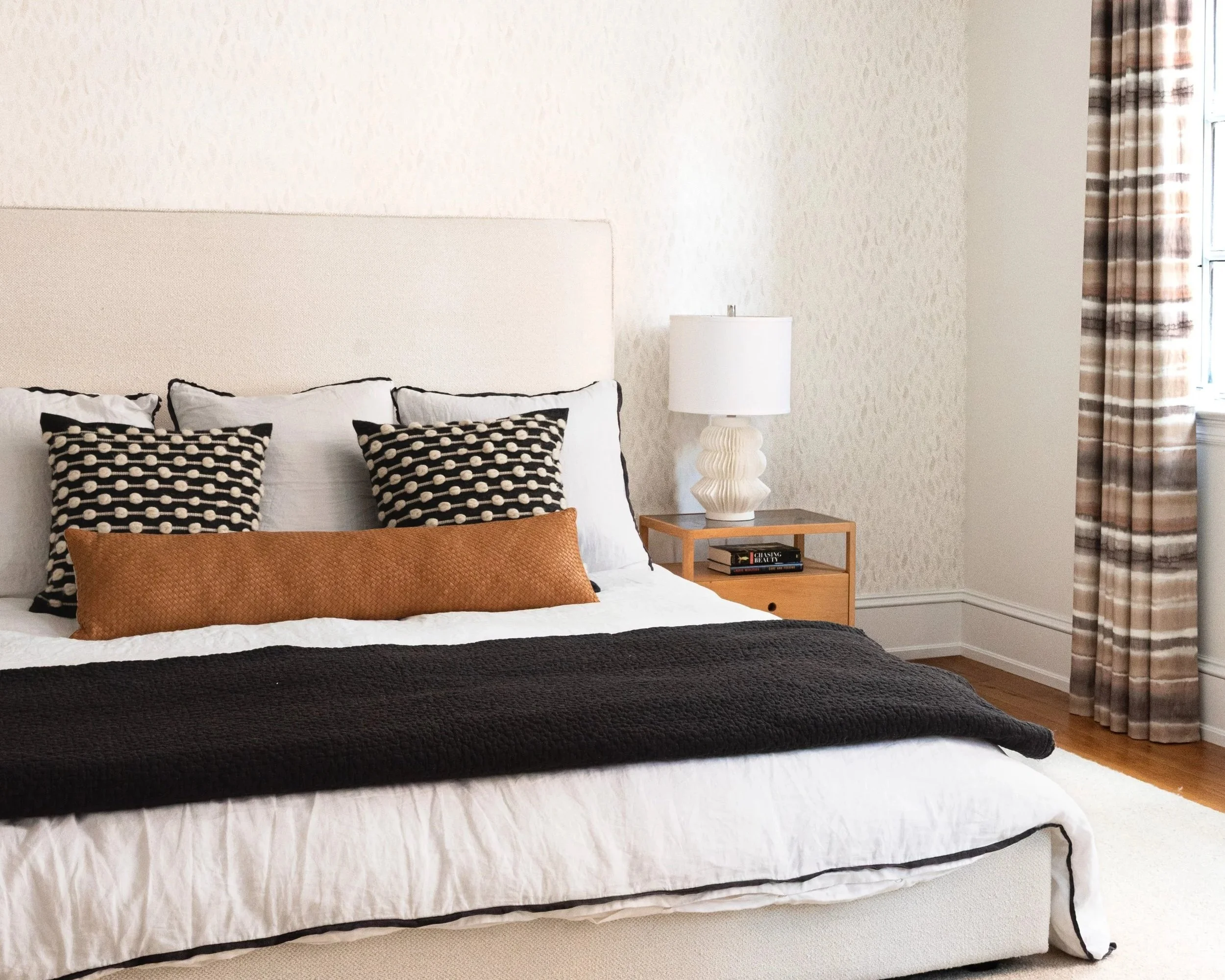 A neatly made bed with white linens, decorative pillows, and a black throw blanket, in a bedroom with a nightstand, white lamp, books, and window with plaid curtains.