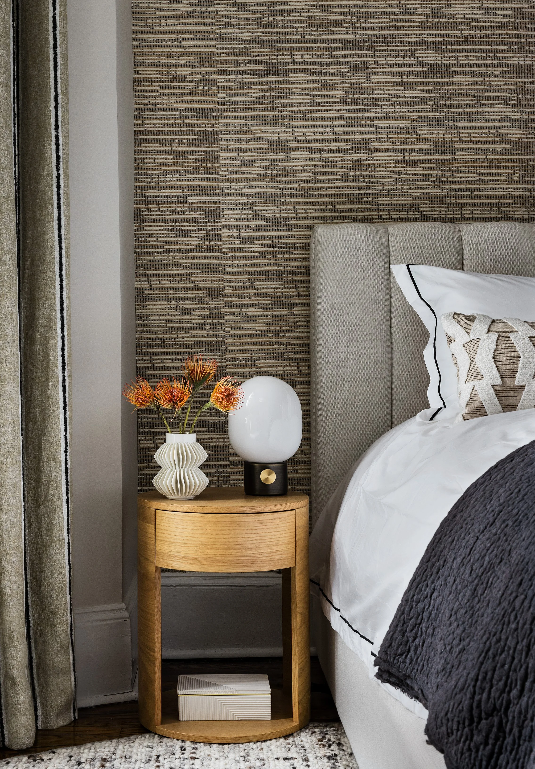 A contemporary bedroom corner featuring a bedside table with a sculptural white vase with orange flowers and a modern lamp, next to a bed with white bedding and decorative pillows, against a textured woven wall and beige curtains.