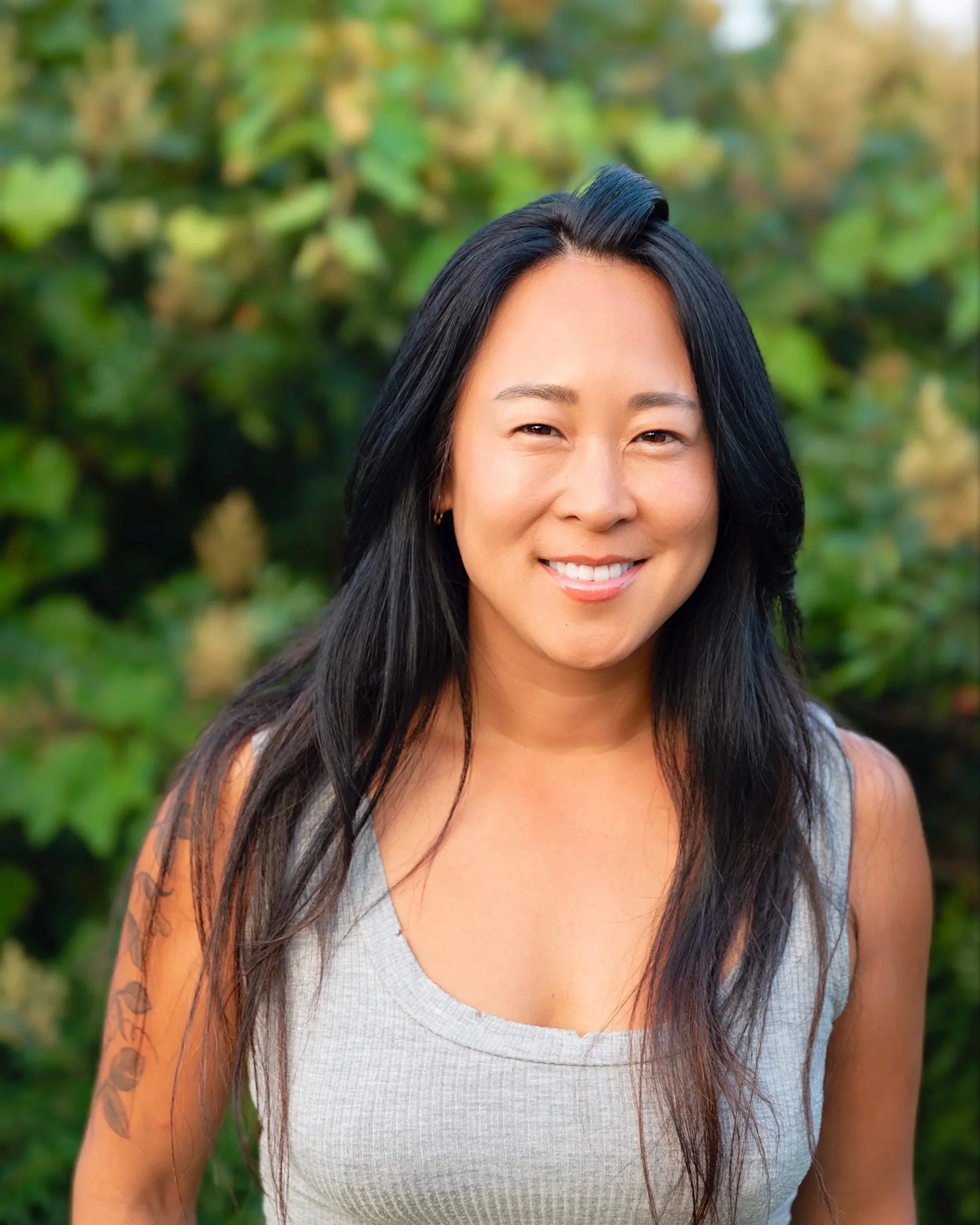 Portrait of a smiling woman with long black hair outdoors, green foliage in the background.