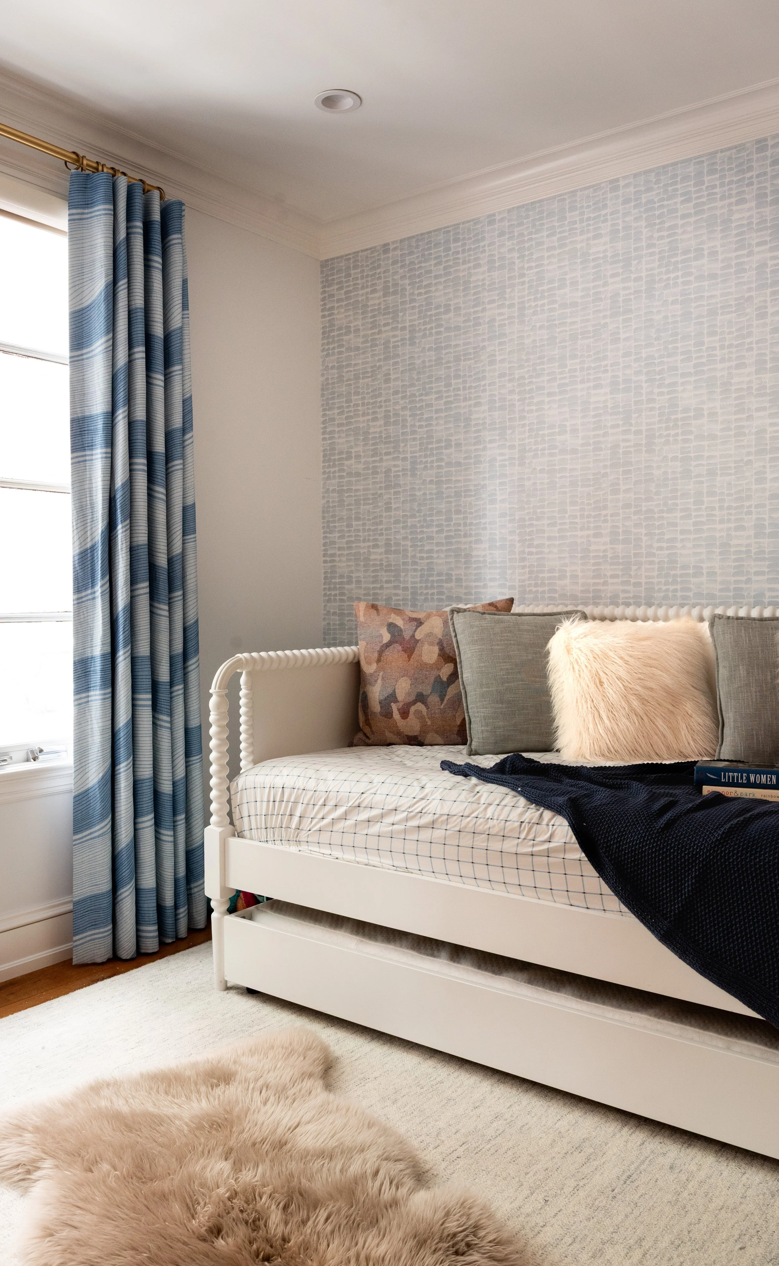 A cozy bedroom corner with a white daybed, decorative pillows, a sheepskin rug, a window with blue patterned curtains, and a textured white wall.