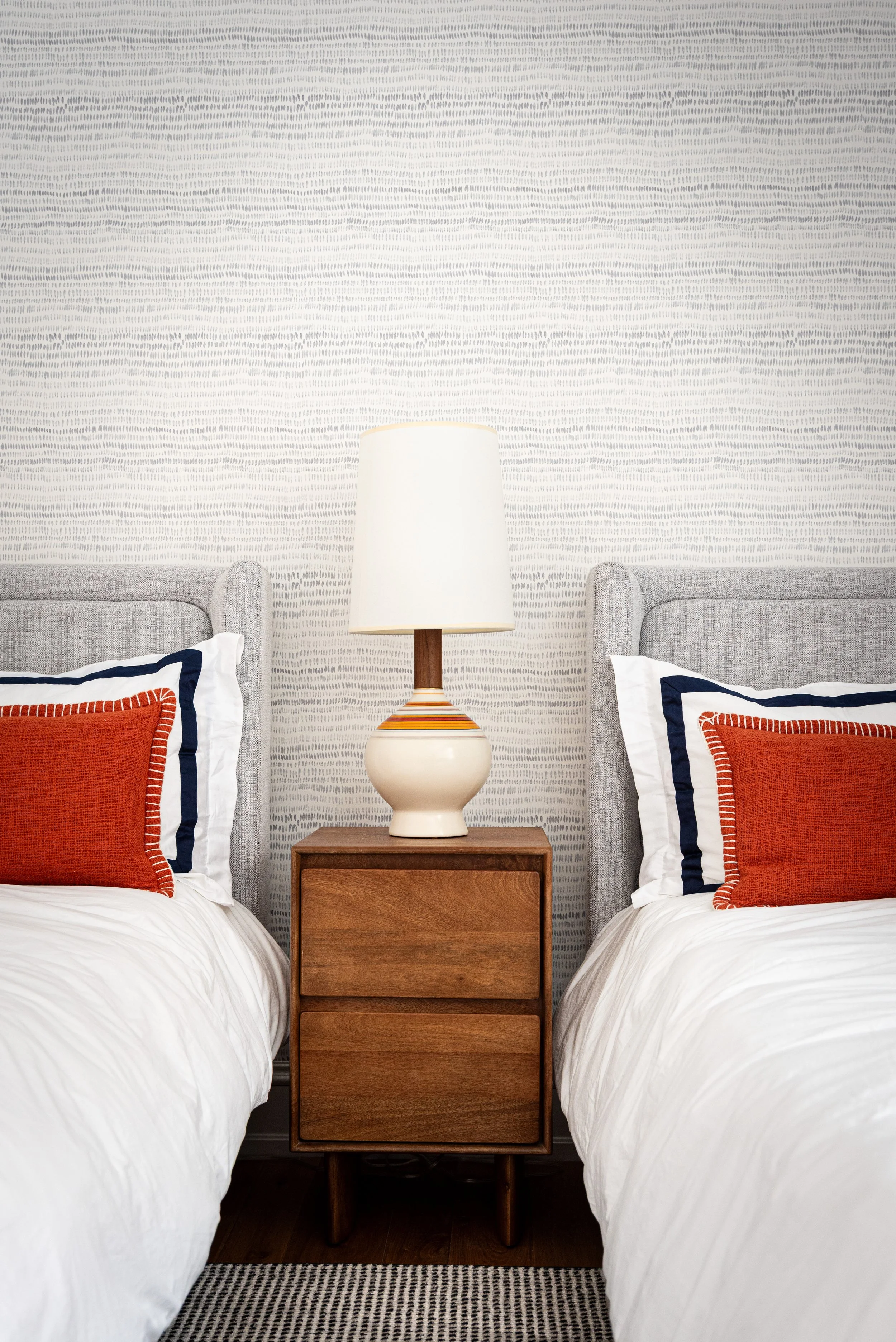 Two beds with matching upholstered headboards separated by a wooden nightstand holding a large table lamp, with decorative pillows and a textured wall in the background.