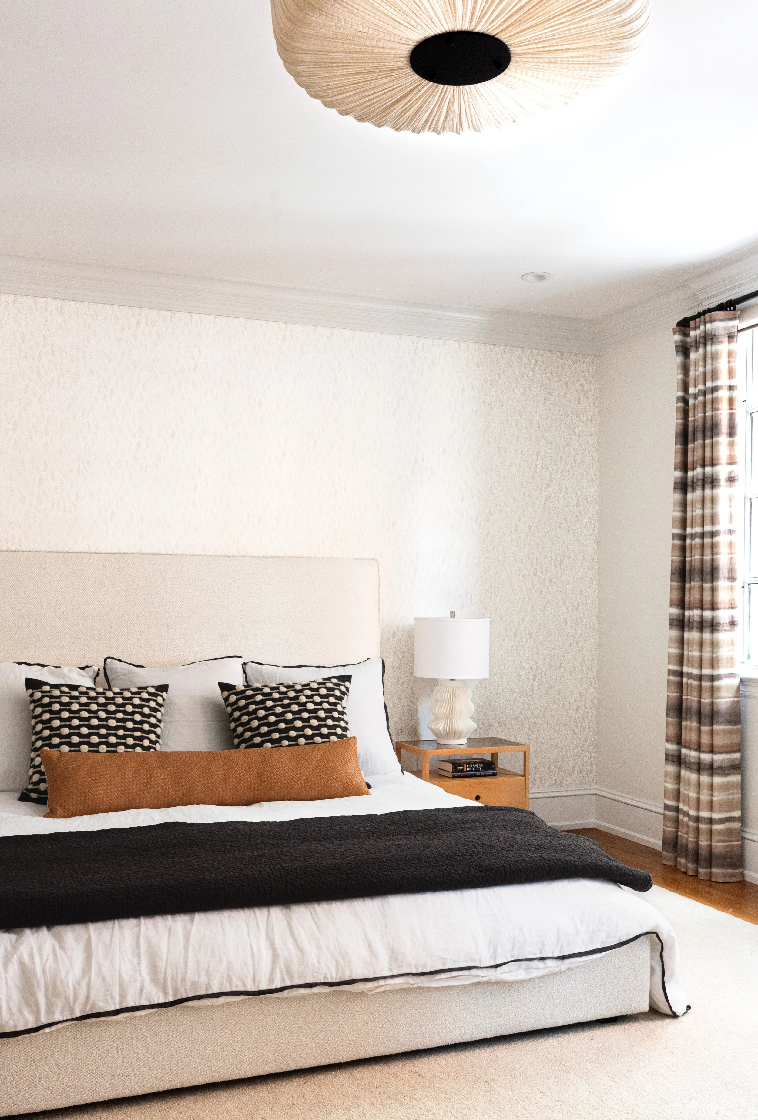 A bedroom with a beige upholstered bed, black and white patterned pillows, a brown bolster pillow, a white bedspread with black trim, and a black throw blanket. There's a wooden nightstand with books and a sculptural white lamp, and a window with pla