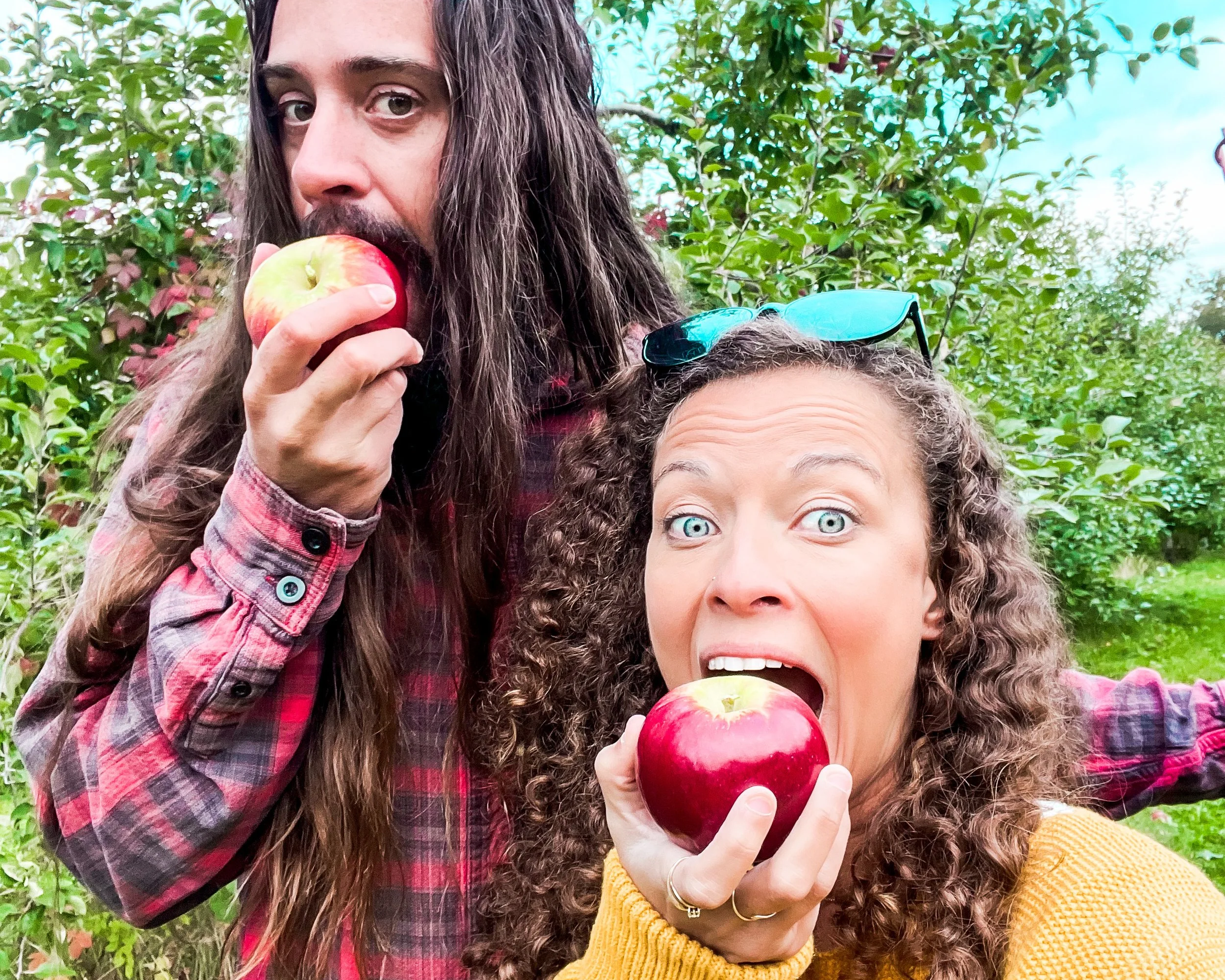 Man with long hair and red flannel next to a woman with curly hair stand in an orchard both taking a bite out of an apple